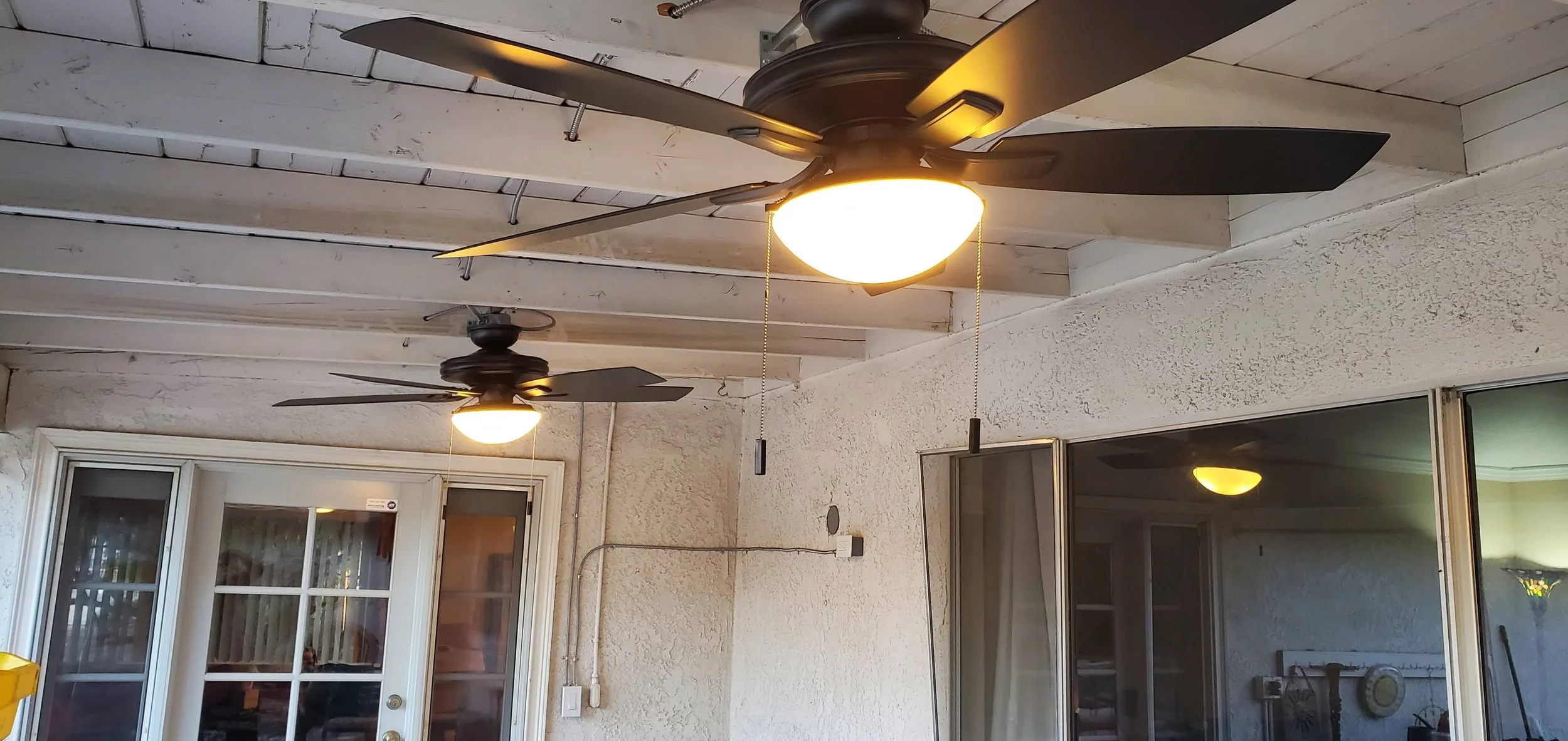 Two ceiling fans with lights on a patio ceiling, with a sliding glass door and windows in the background.