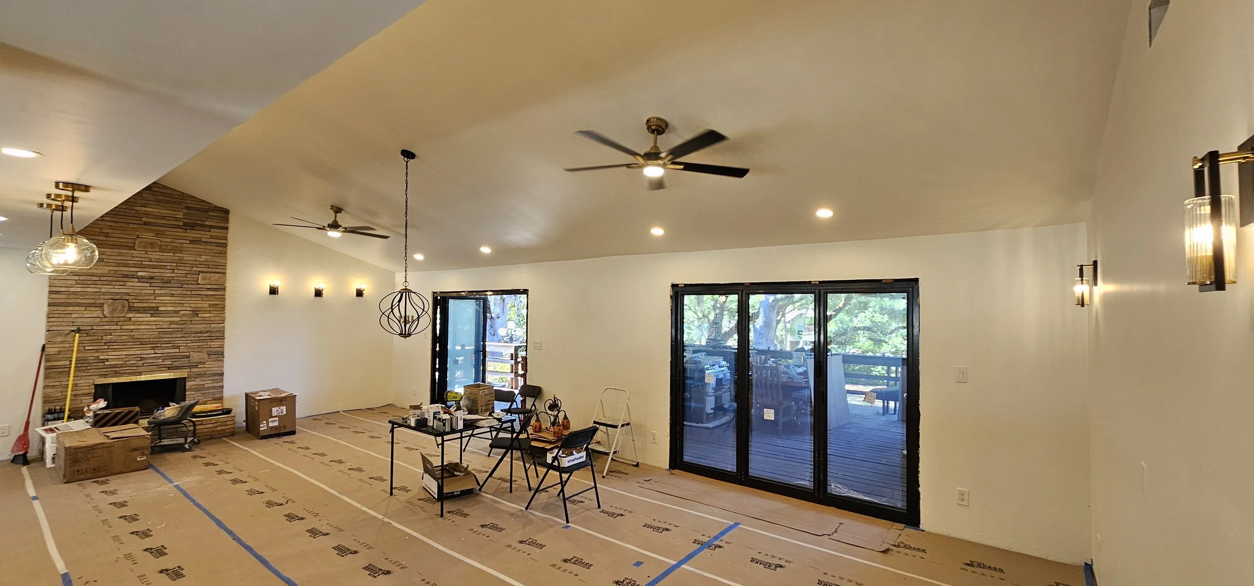 Living room under renovation with cardboard on floor, construction tools and furniture, ceiling fans, and sliding glass doors leading to a deck outside.