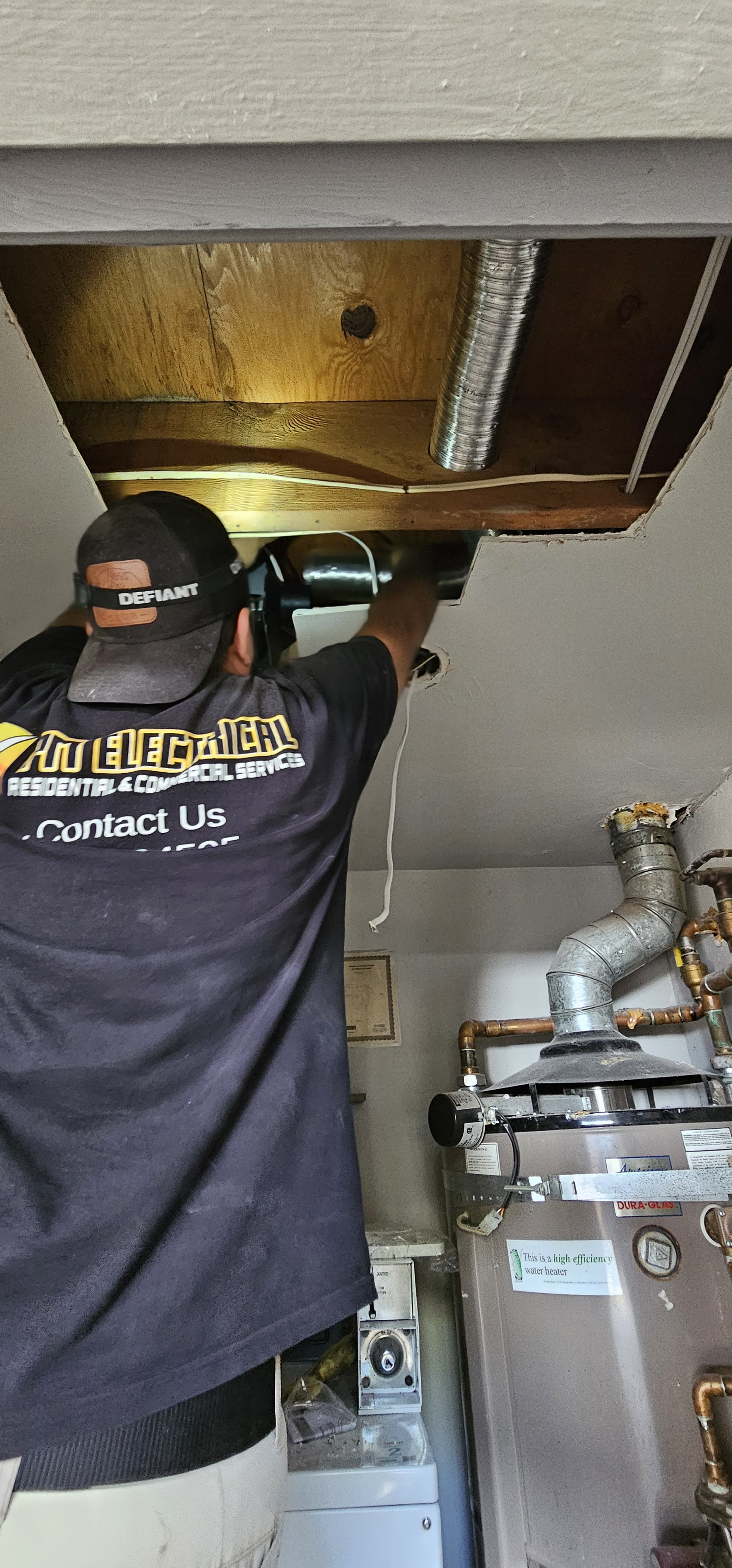 A worker in black clothing and a cap labeled 'Defiant' is repairing or installing a vent or duct in a ceiling. The work area contains pipes, a water heater, and a washing machine underneath.