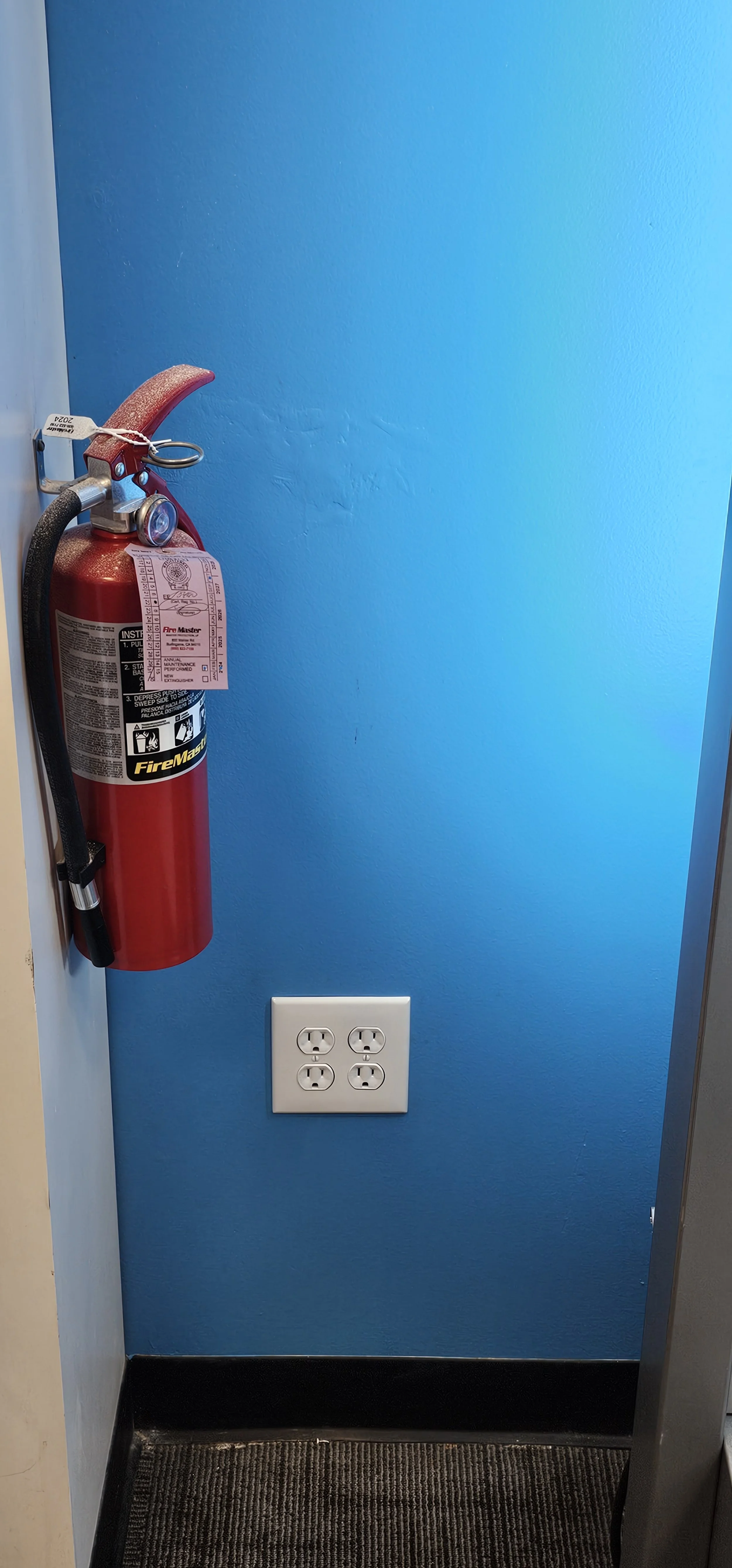 A fire extinguisher mounted on a blue wall next to an electrical outlet.