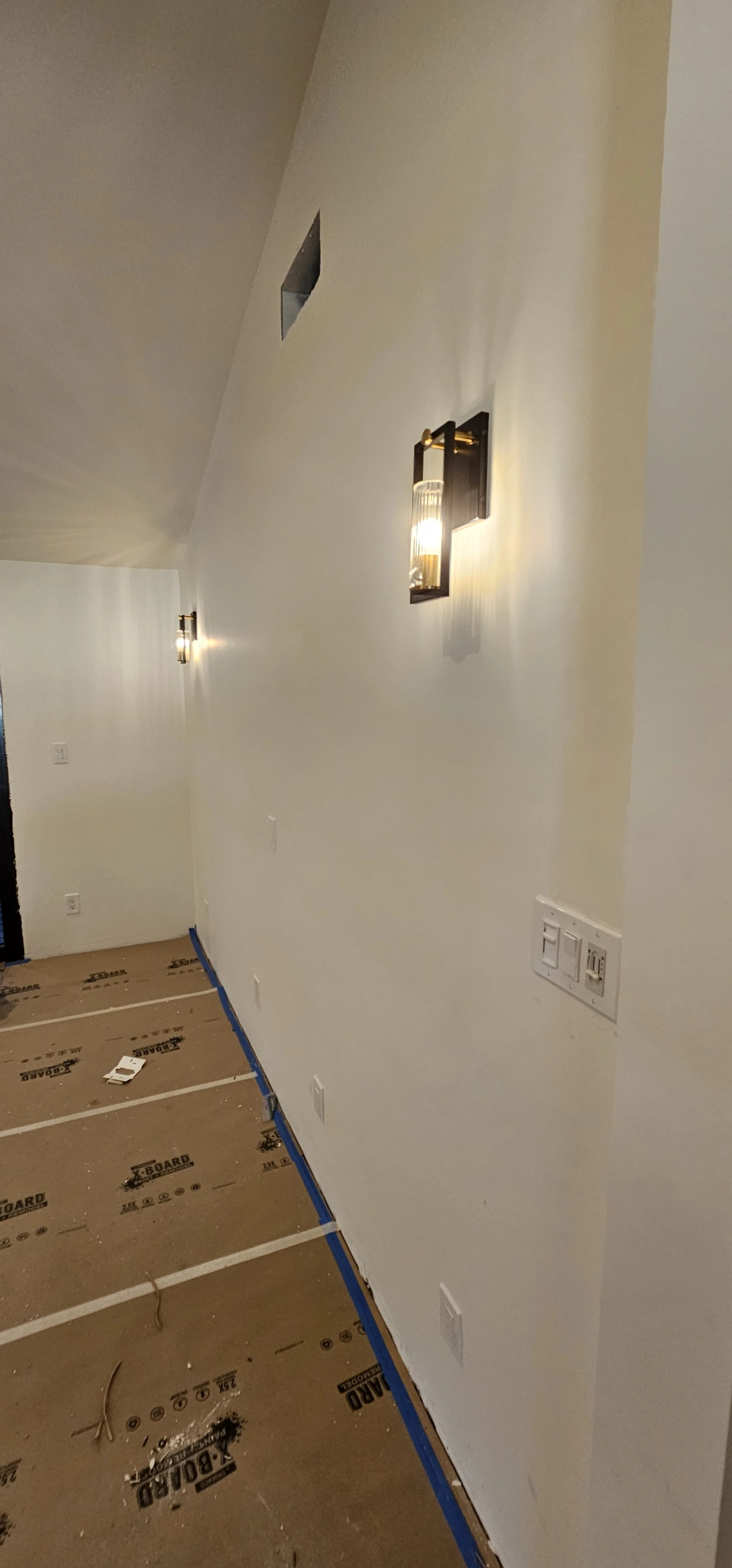 A white interior wall with two modern wall sconces emitting warm light and a small rectangular opening near the ceiling. The floor is covered with brown construction paper with black markings and blue painter's tape.