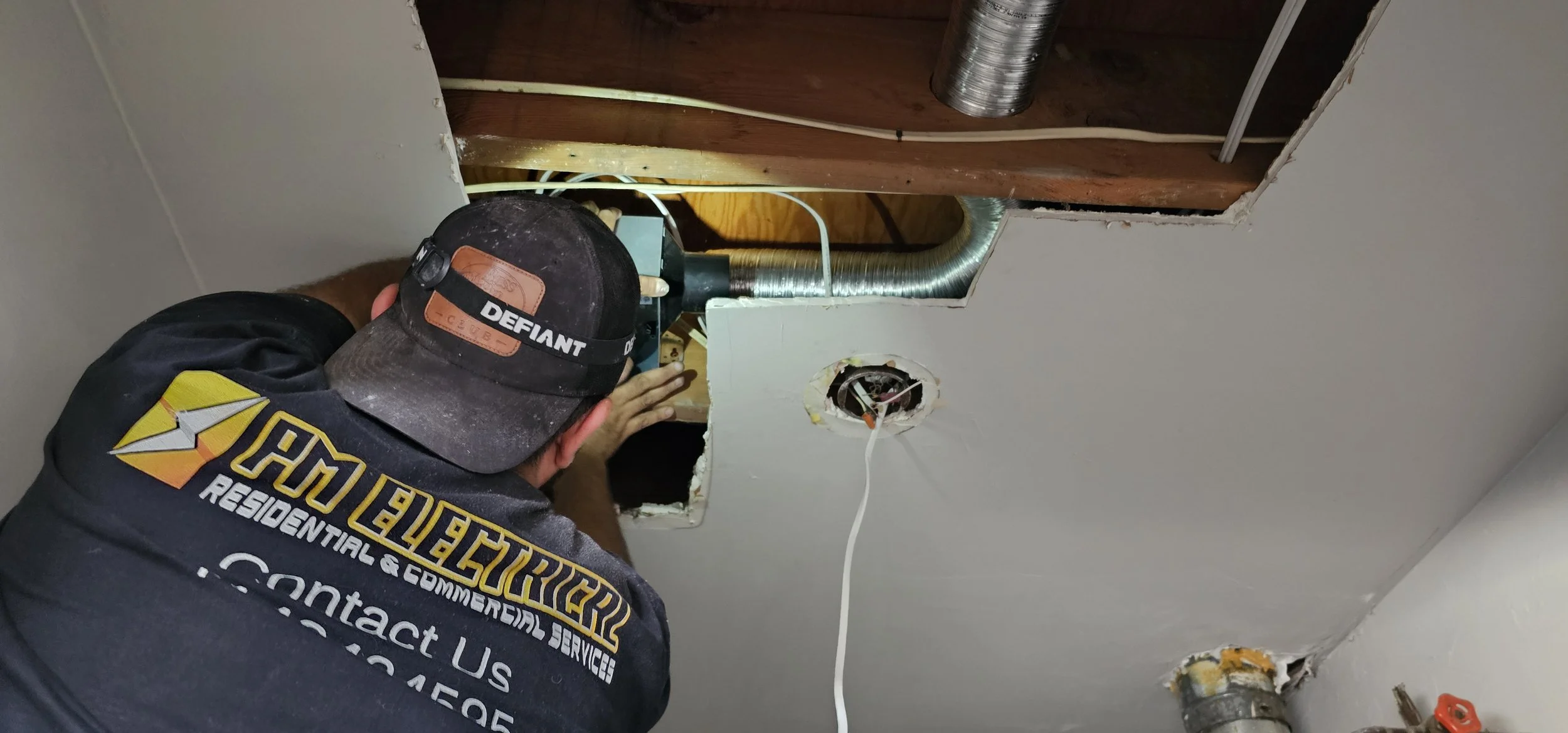 A technician working on electrical wiring inside a ceiling opening, with exposed ductwork and a light fixture hole nearby.