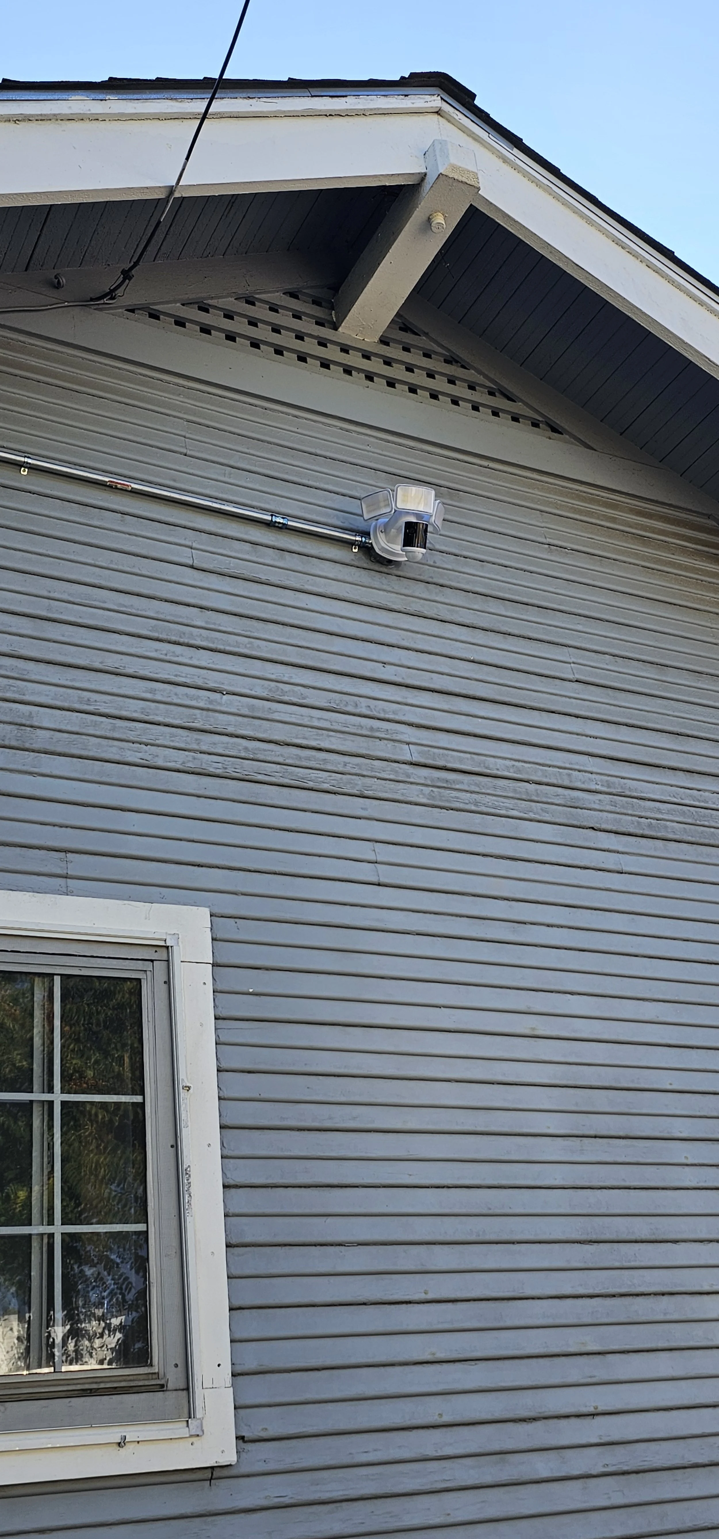 Close-up of the exterior of a house showing gray horizontal siding, a window with white trim, and a security camera mounted on the wall, with part of the roof and blue sky visible.