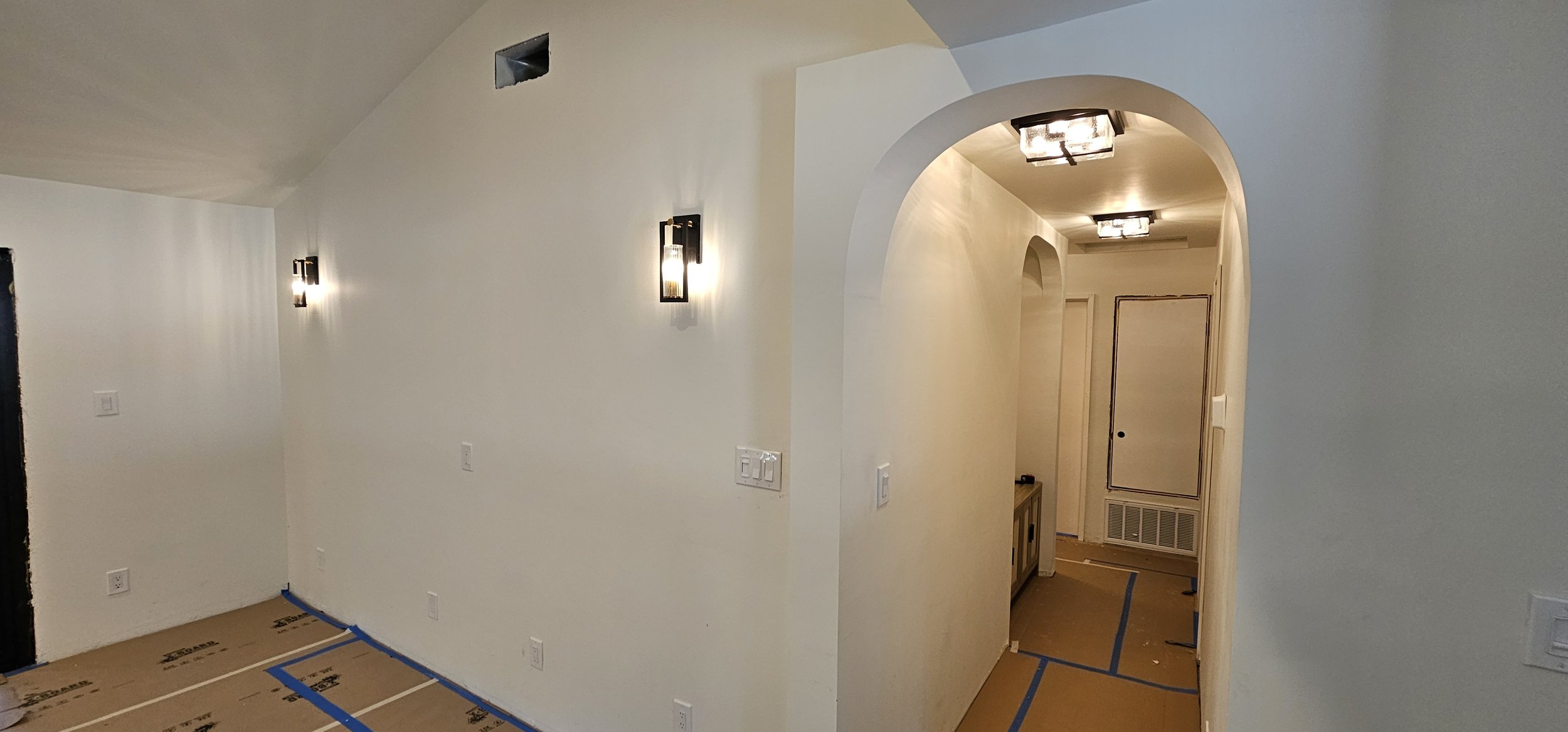 Interior of a house under construction with white walls, decorative wall sconces, and a hallway with arched doorways, showing protective brown paper on the floor.
