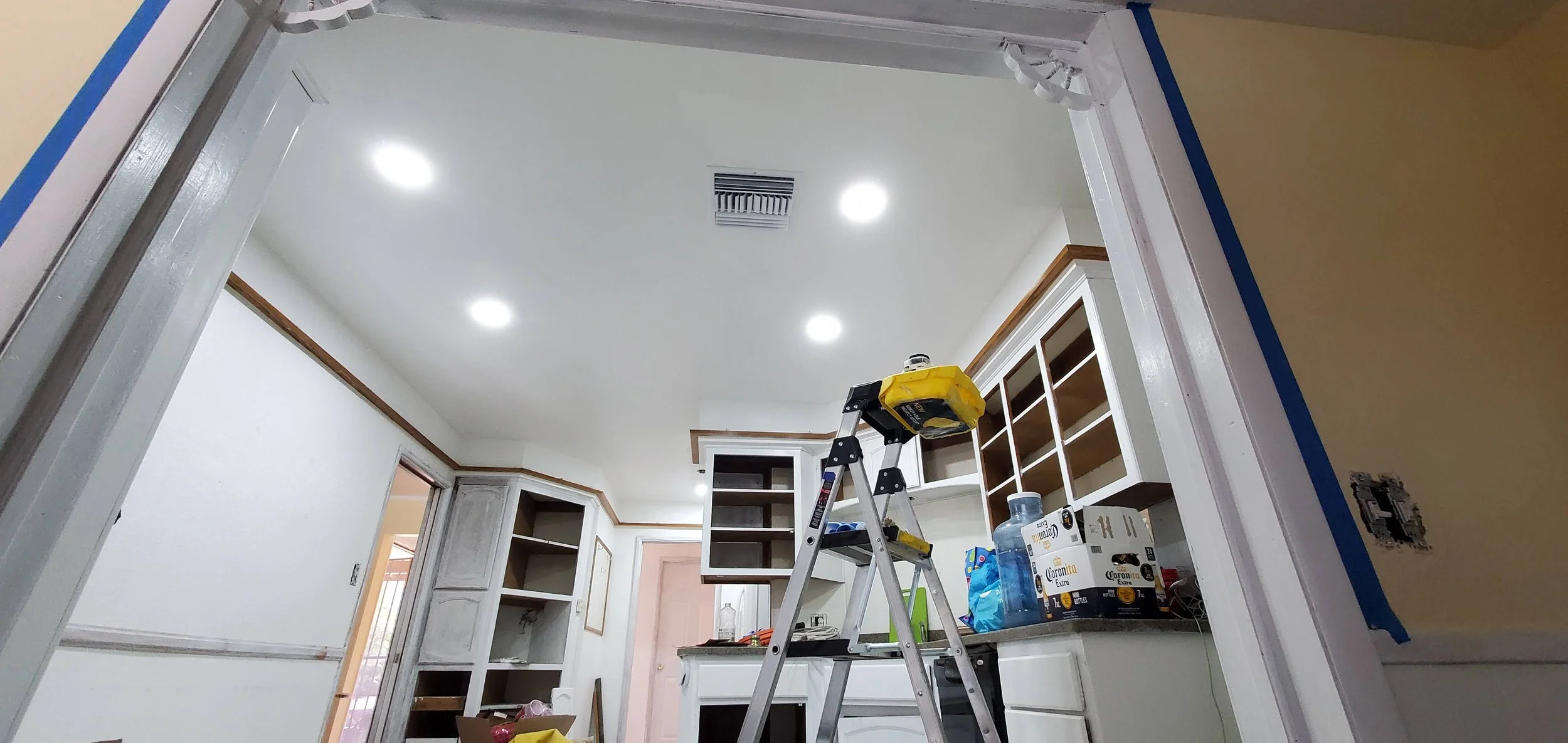 Interior of a kitchen under renovation with an aluminum ladder, some unopened boxes of Corona beer, bottles, and construction supplies on the countertops, and open cabinets on the wall.