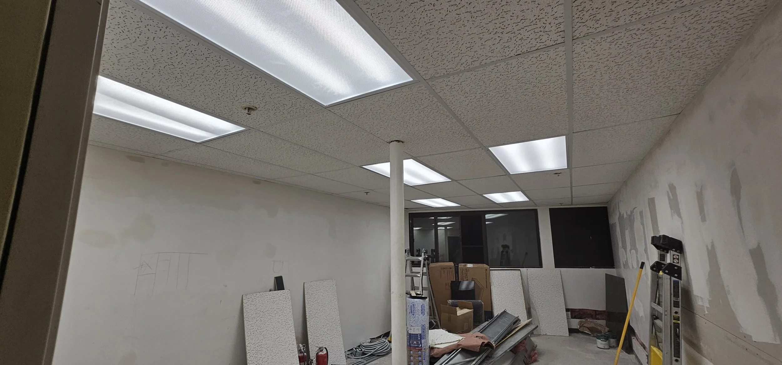 Interior of a room under renovation with ceiling lights, construction materials, and tools, including stairs, ladders, and drywall panels.