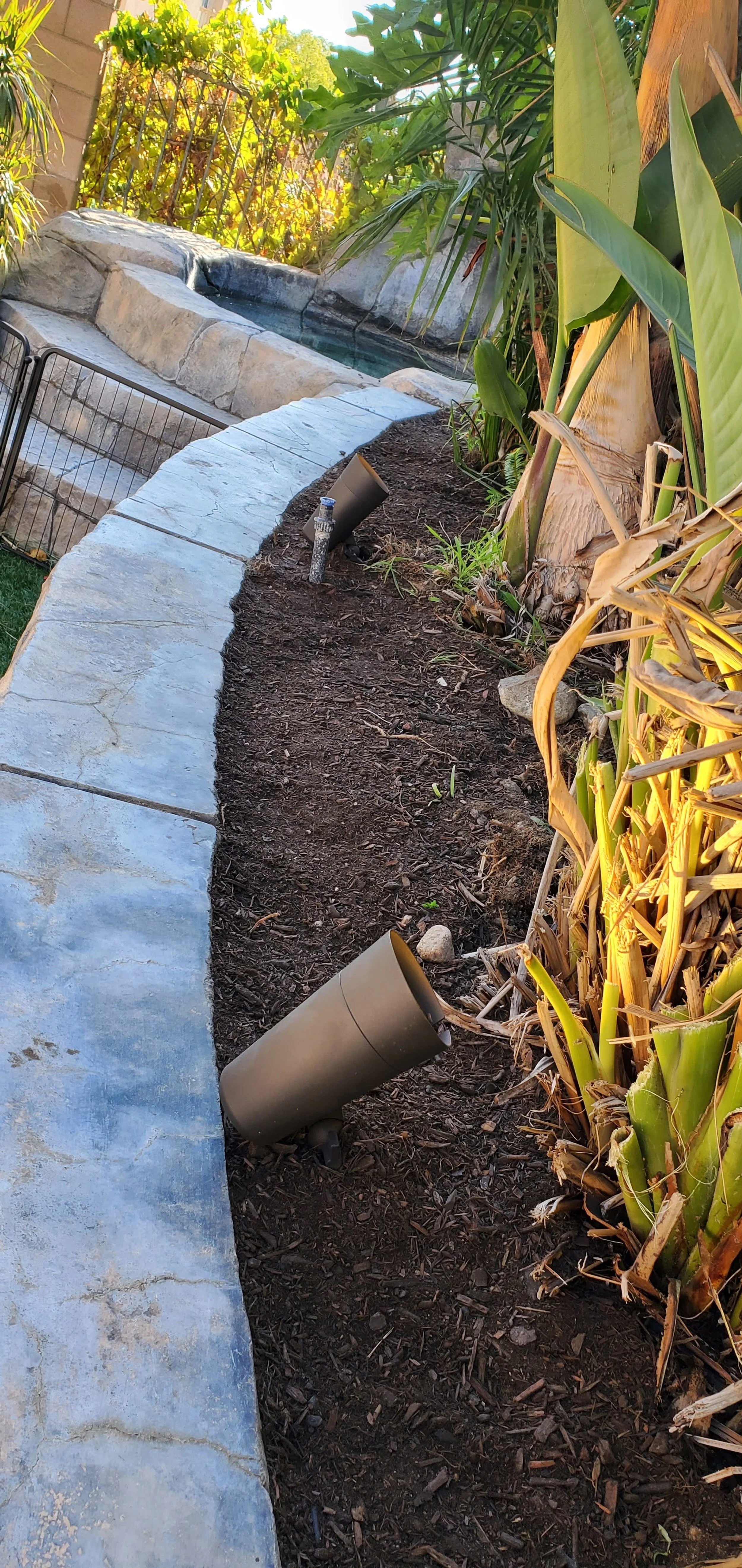 A garden bed with soil and plants next to a stone pathway, with outdoor landscape lights and a small hot tub in the background.