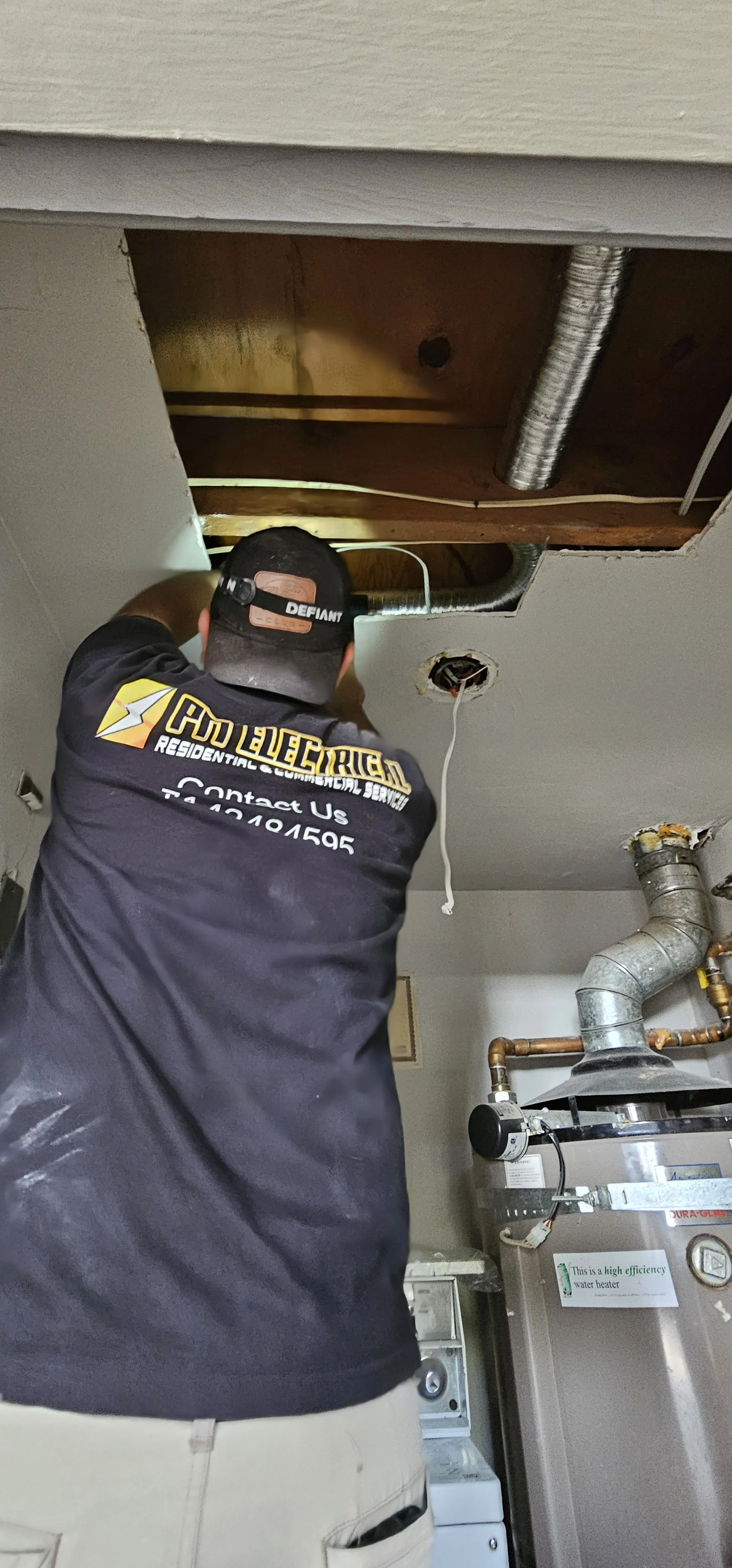 A technician working on a ceiling, with a hole cut out revealing ductwork and wiring, in a utility room with water heater and washer.