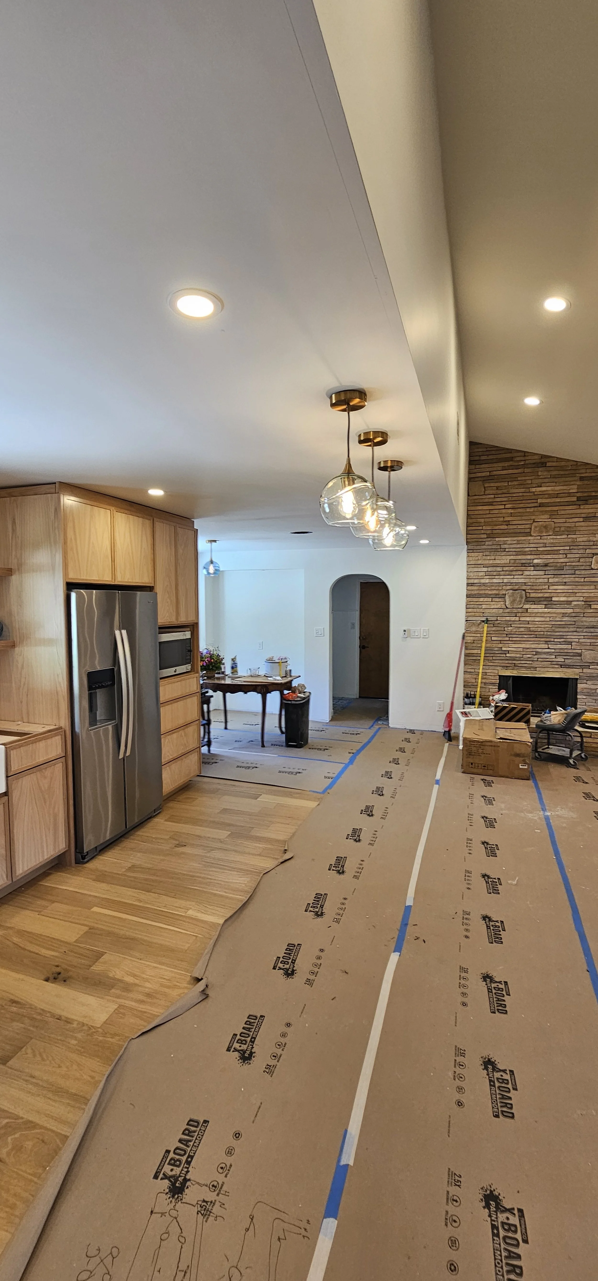 Living room undergoing renovation with partially covered floor, kitchen with wooden cabinets and stainless steel refrigerator, and brick fireplace. Construction tools and materials are present.