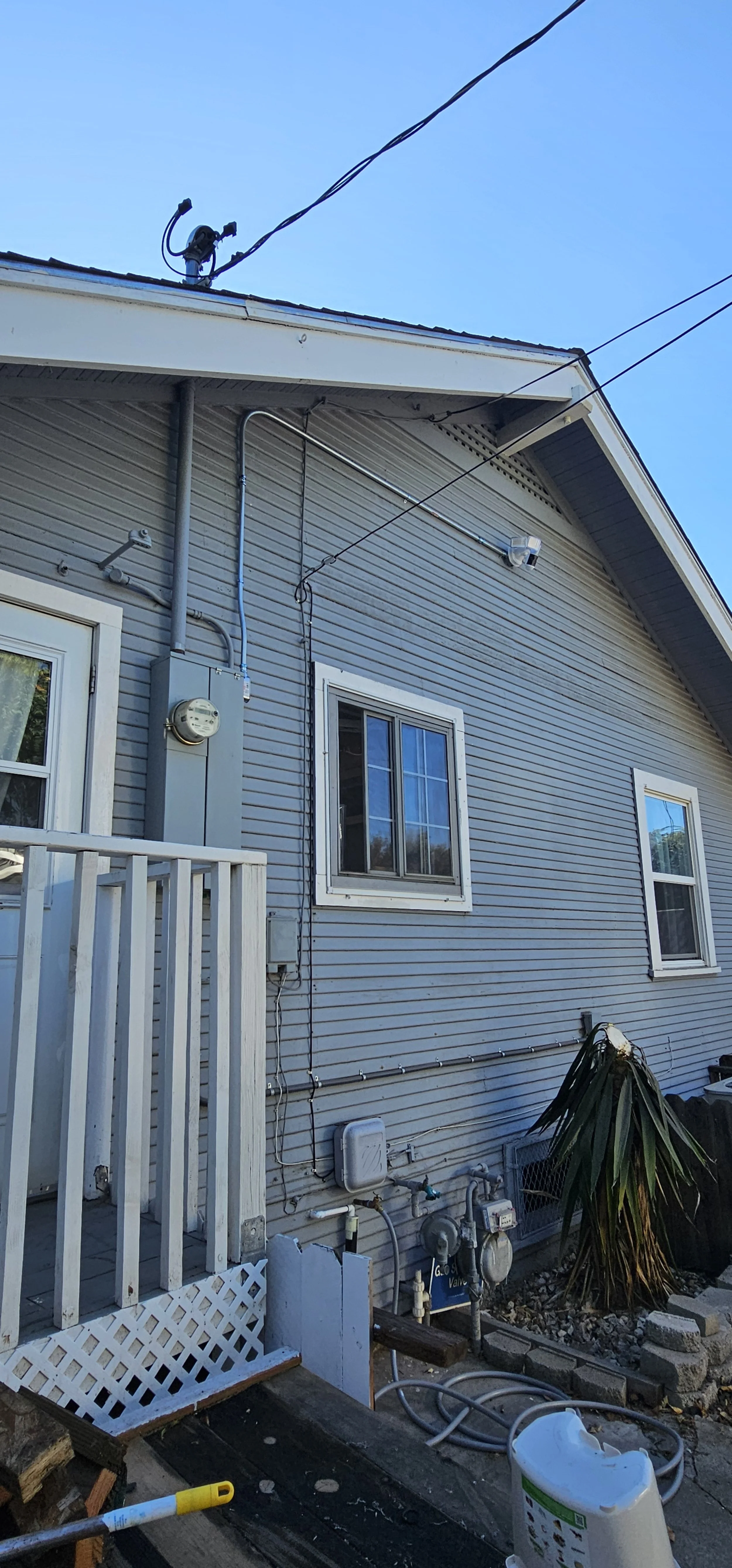 Side view of a house exterior showing two windows, utility meters, wiring, and a small garden with a plant.