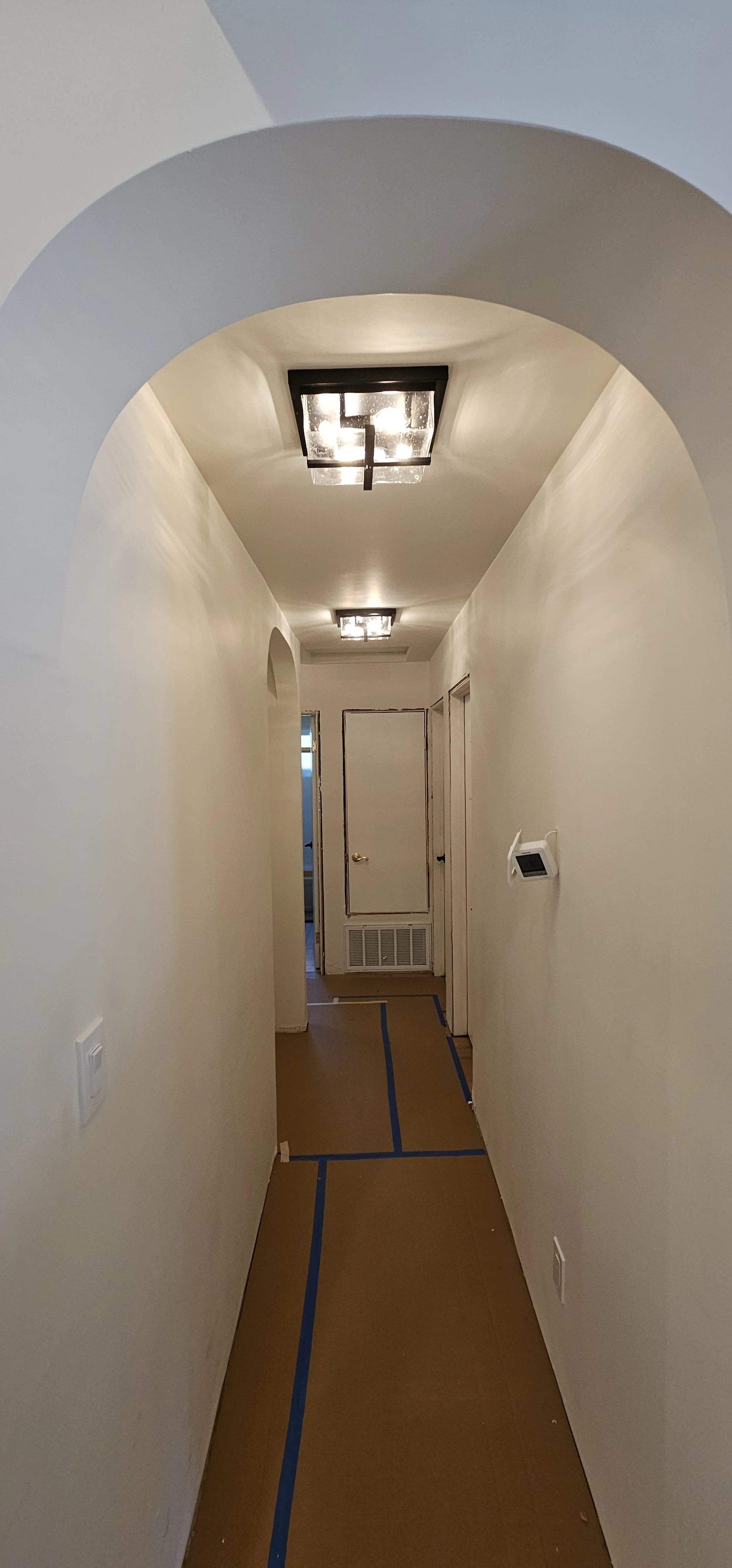A narrow hallway with beige painted walls, brown protective floor covering taped with blue painter's tape, ceiling lights, a thermostat on the right wall, and a door at the end of the hallway.