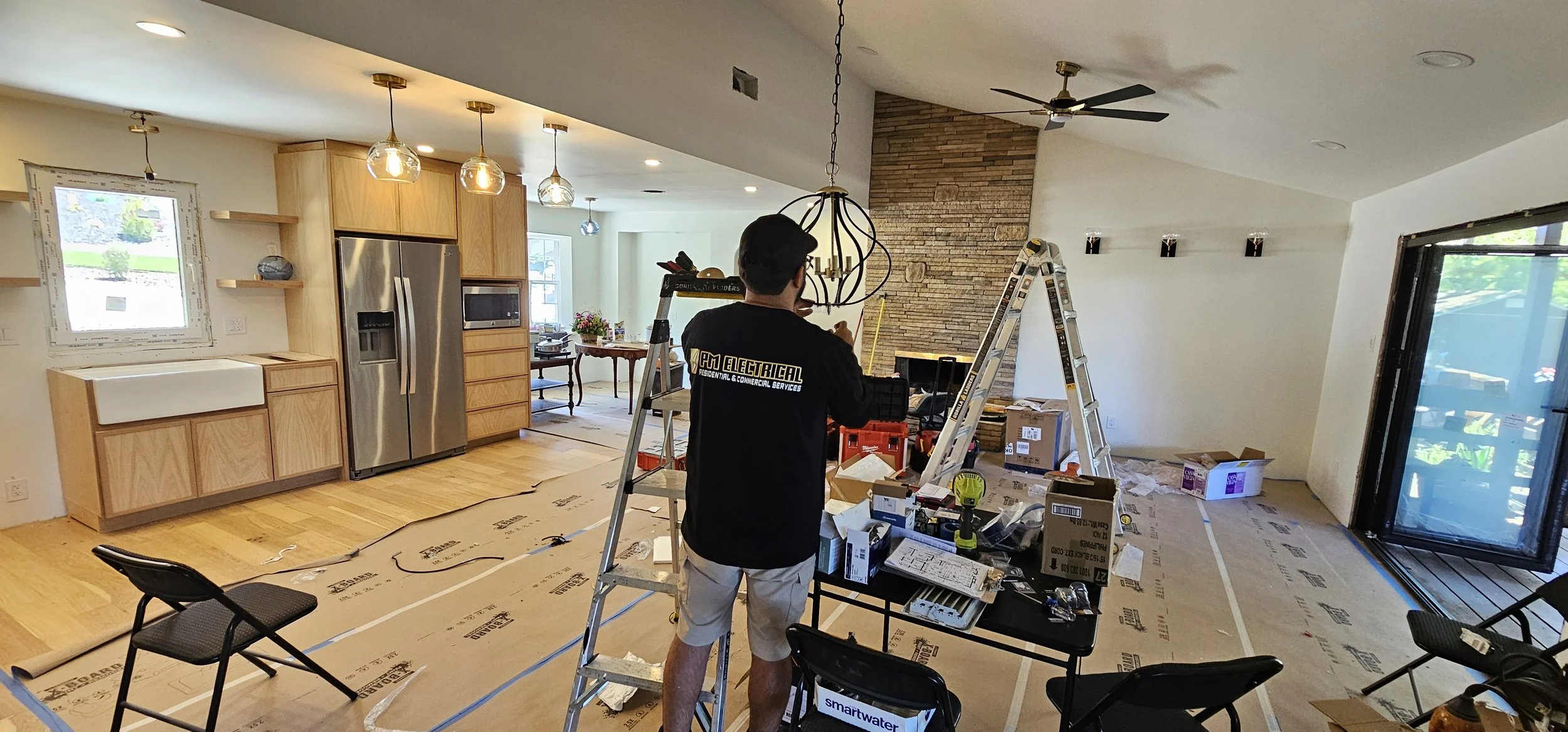 Interior of a house under renovation with a man working on electrical wiring, surrounded by ladders, tools, and boxes, with a kitchen on the left and a brick fireplace on the right.