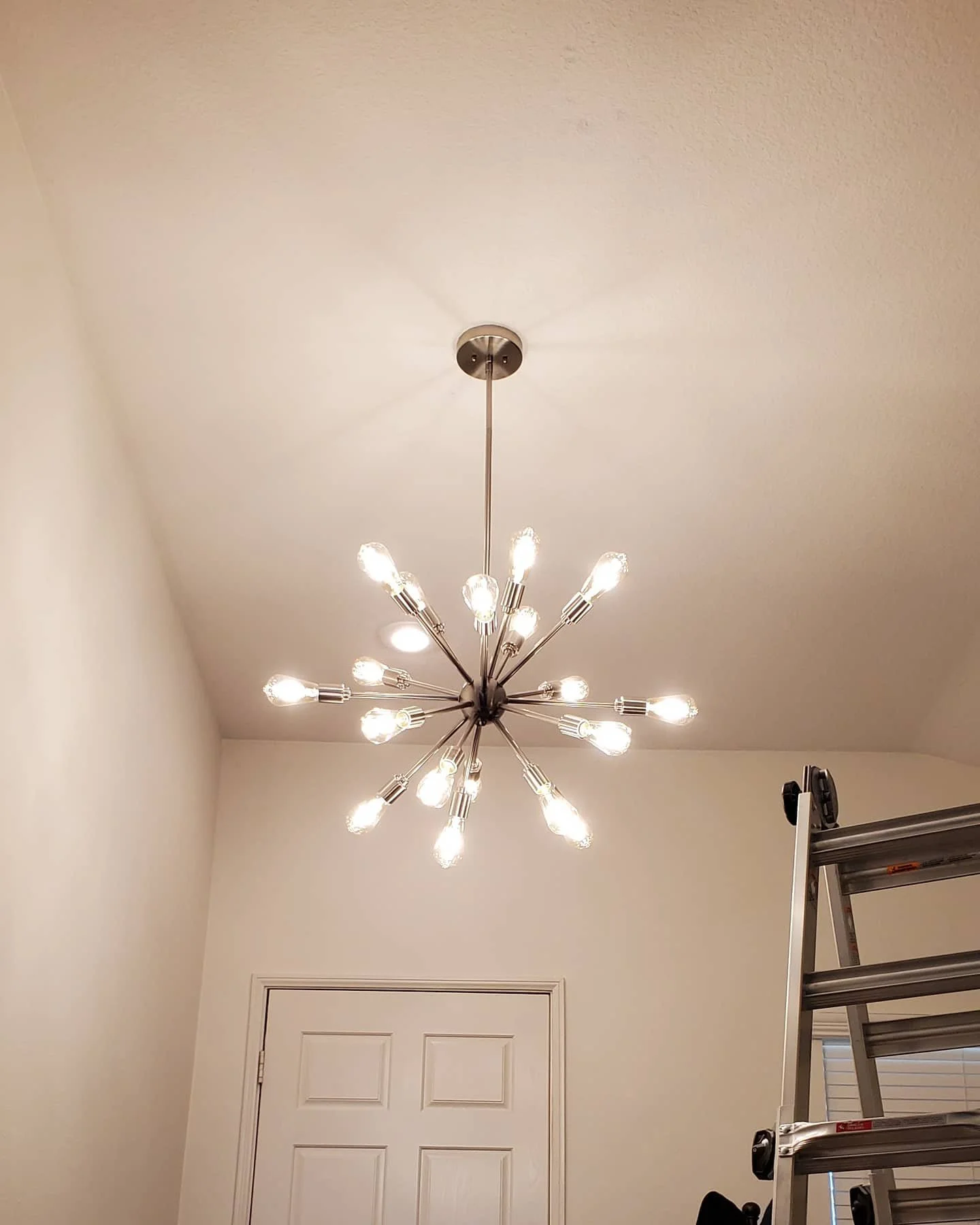 Modern chandelier with multiple exposed bulbs hanging from the ceiling in a room with a closed door and a ladder.