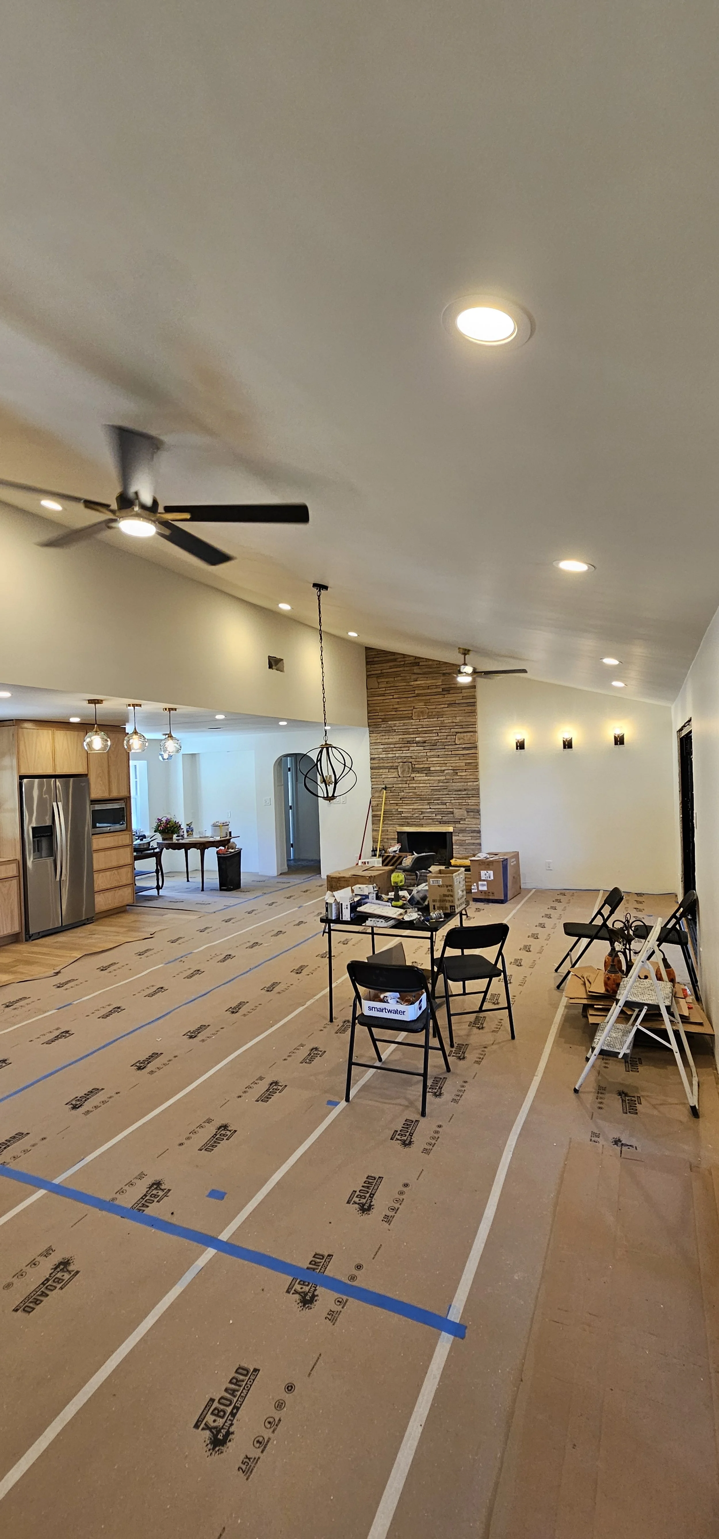 Living room under construction with temporary floor covering, kitchen area with appliances, dining table, and furniture in the background. Tools and boxes are scattered around.