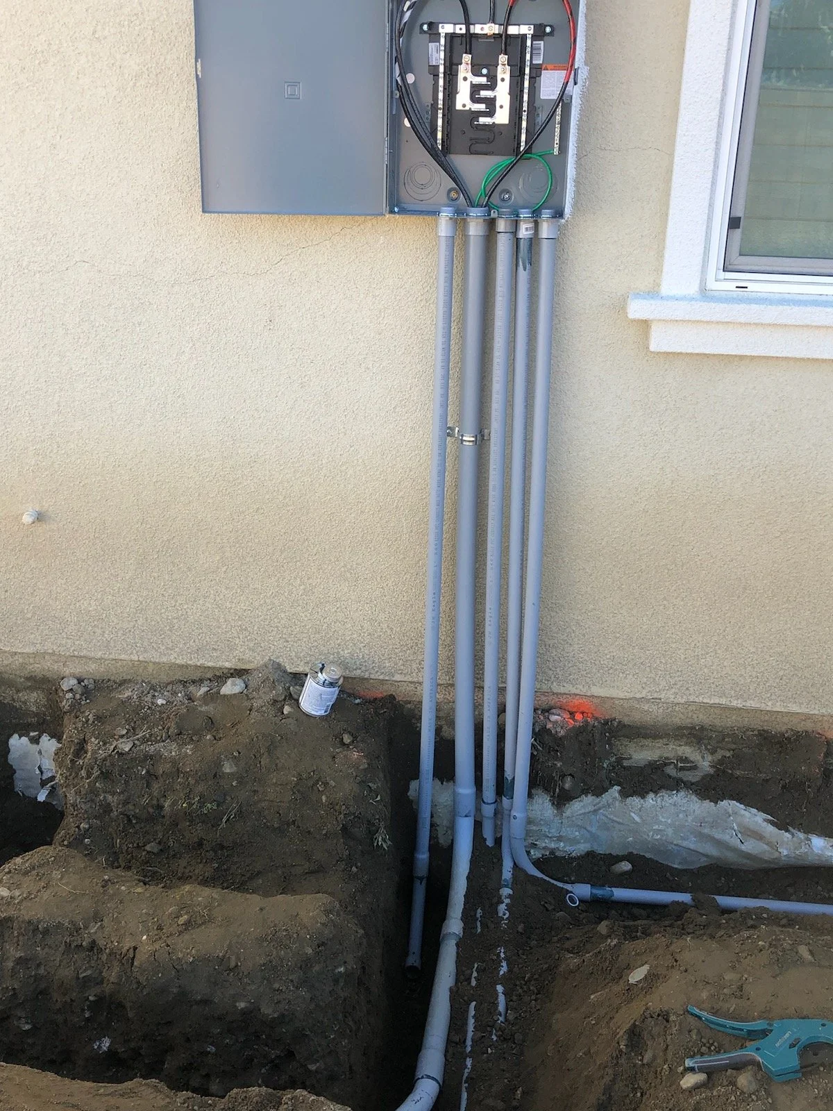 Electrical wiring and pipes installed outside the house near a window, with soil and construction tools in the foreground.
