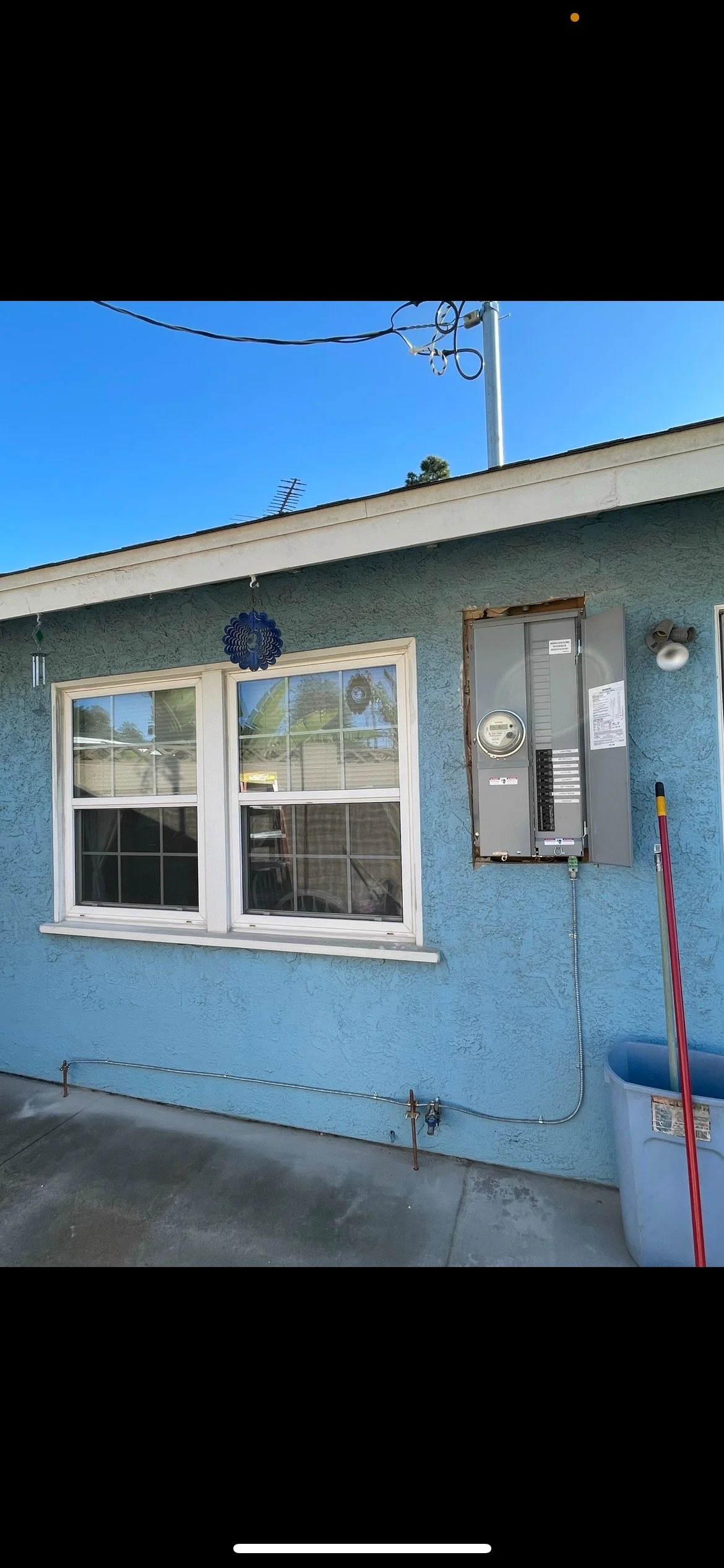 A house wall painted light blue with a white-framed double window. An electrical meter is mounted on the wall next to the window, with an open metal panel cover. There is a red broomstick leaning against the wall near a large gray trash bin. A decorative blue wind chime hangs under the roofline, and a security camera is mounted near the window. Utility wires run across the top of the image. The sky is clear and blue.