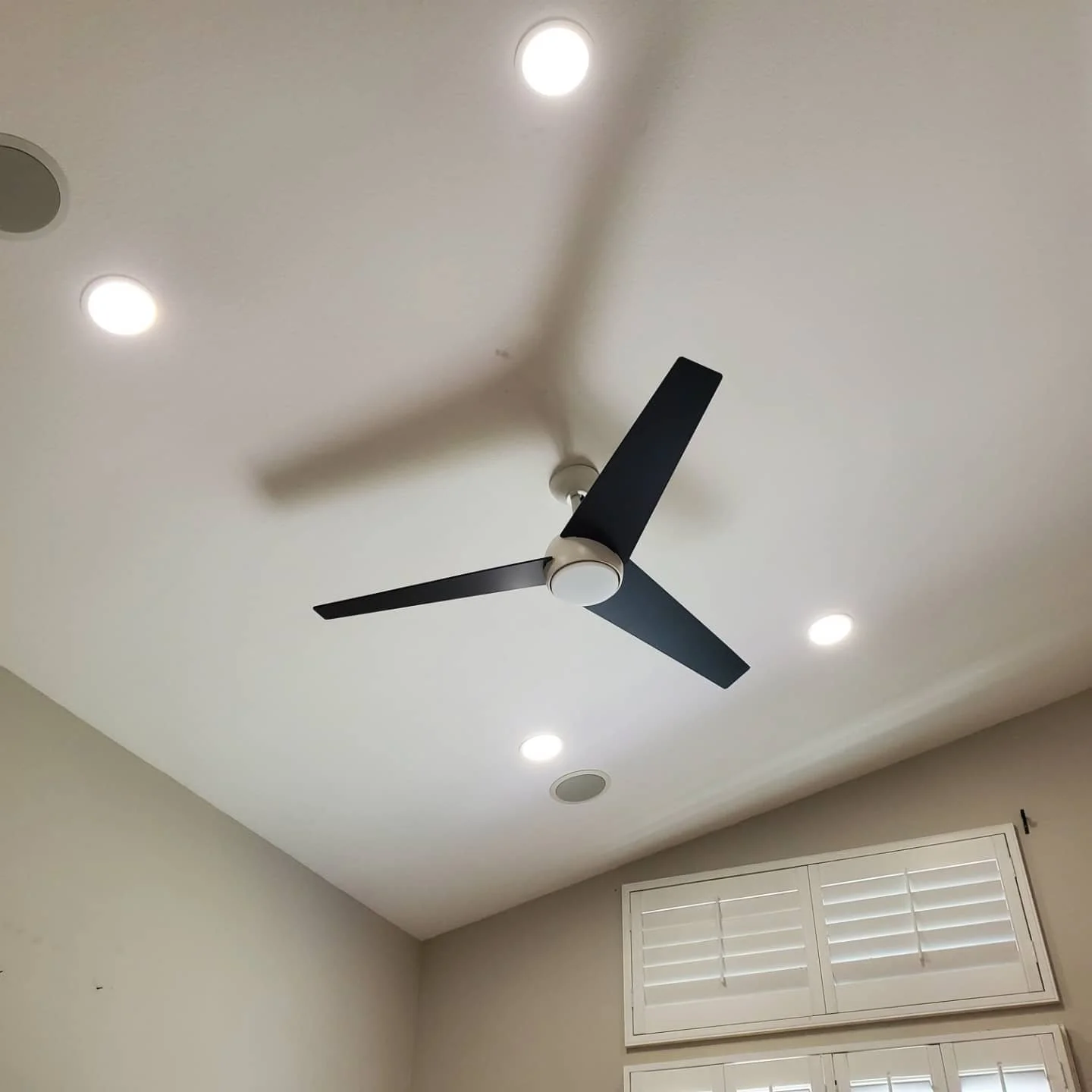 Ceiling fan with three black blades, installed on a white ceiling with four recessed lights, in a room with beige walls and a window with white shutters.