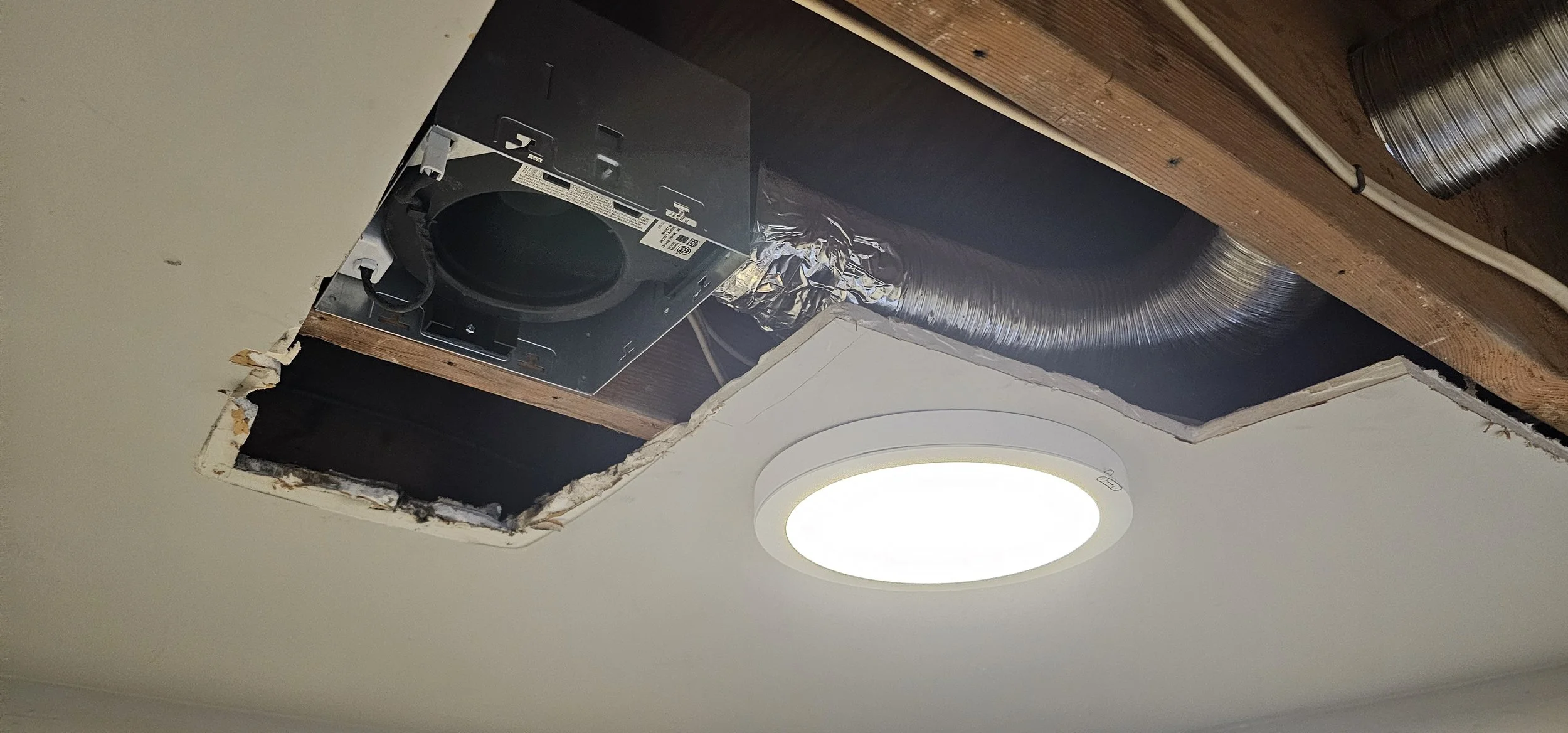 Ceiling with a hole where a light fixture and HVAC duct are being installed. The light is a round LED panel, and the ductwork and wiring are exposed.