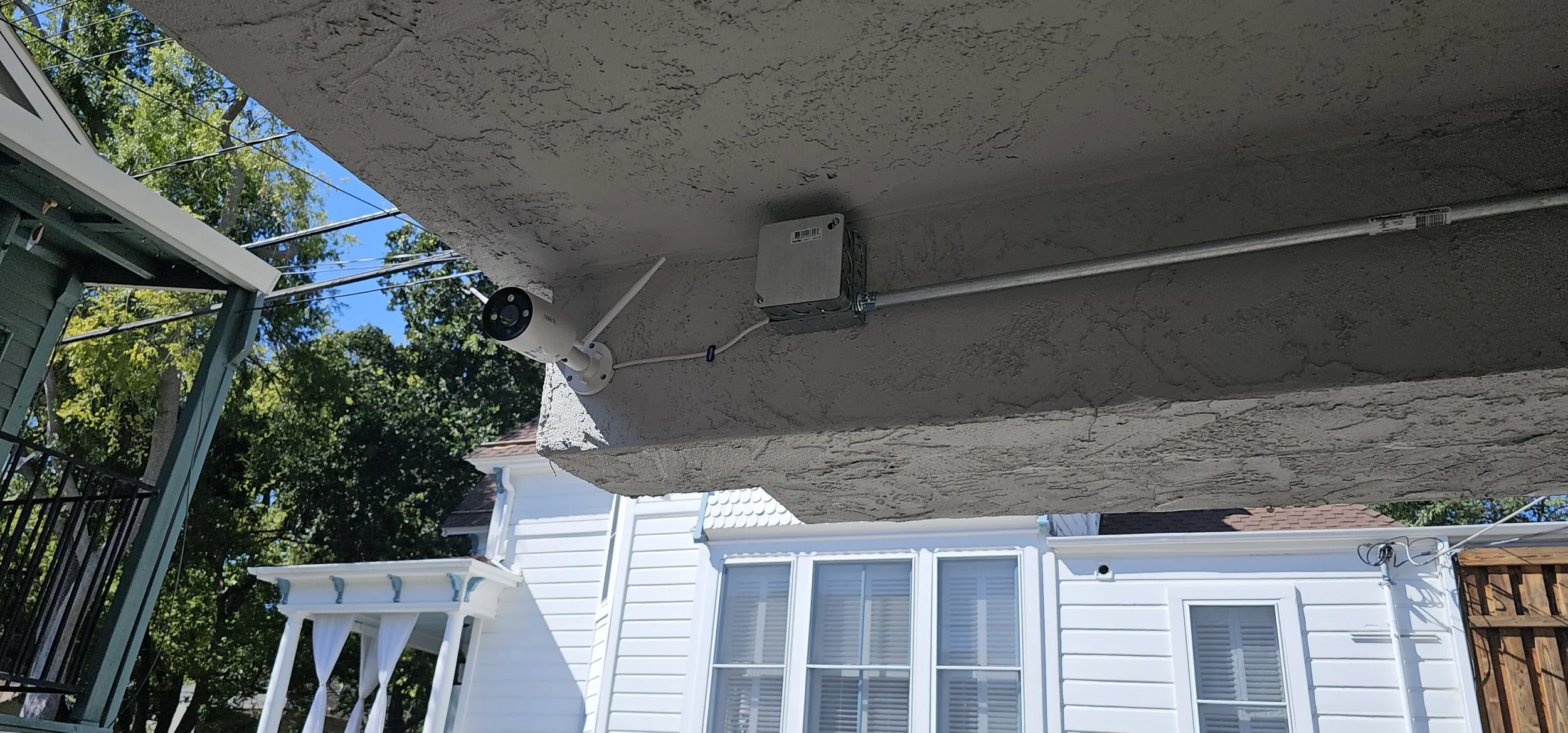 Security camera mounted on a concrete wall with electrical conduit, with houses, trees, and blue sky in the background.