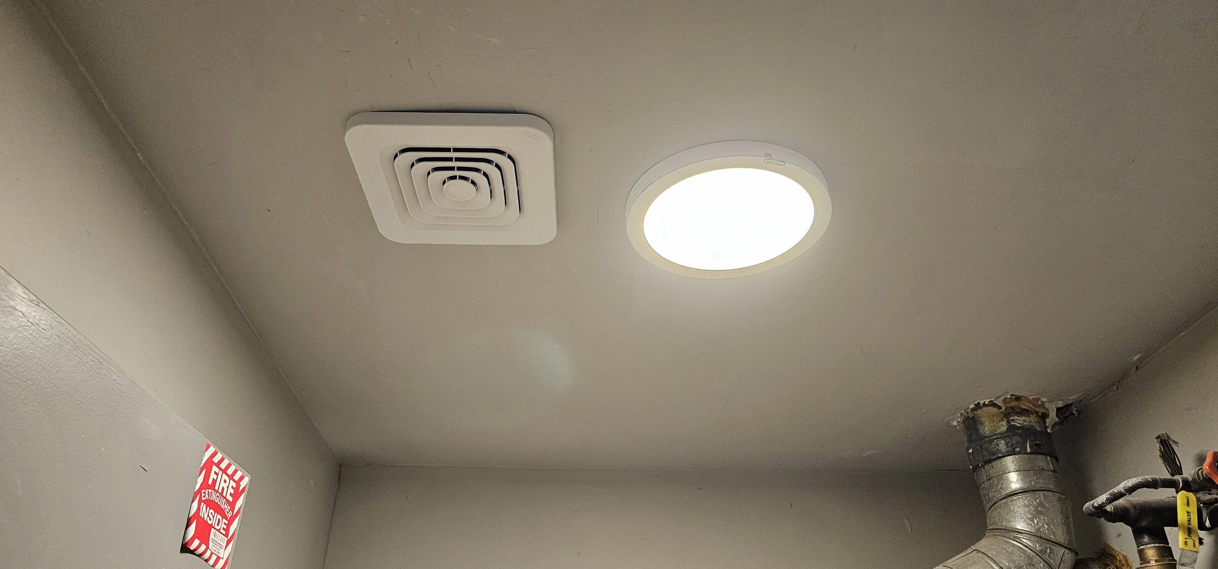 Ceiling with a square exhaust fan, a circular light fixture, a fire extinguisher sign, and a metal duct pipe.