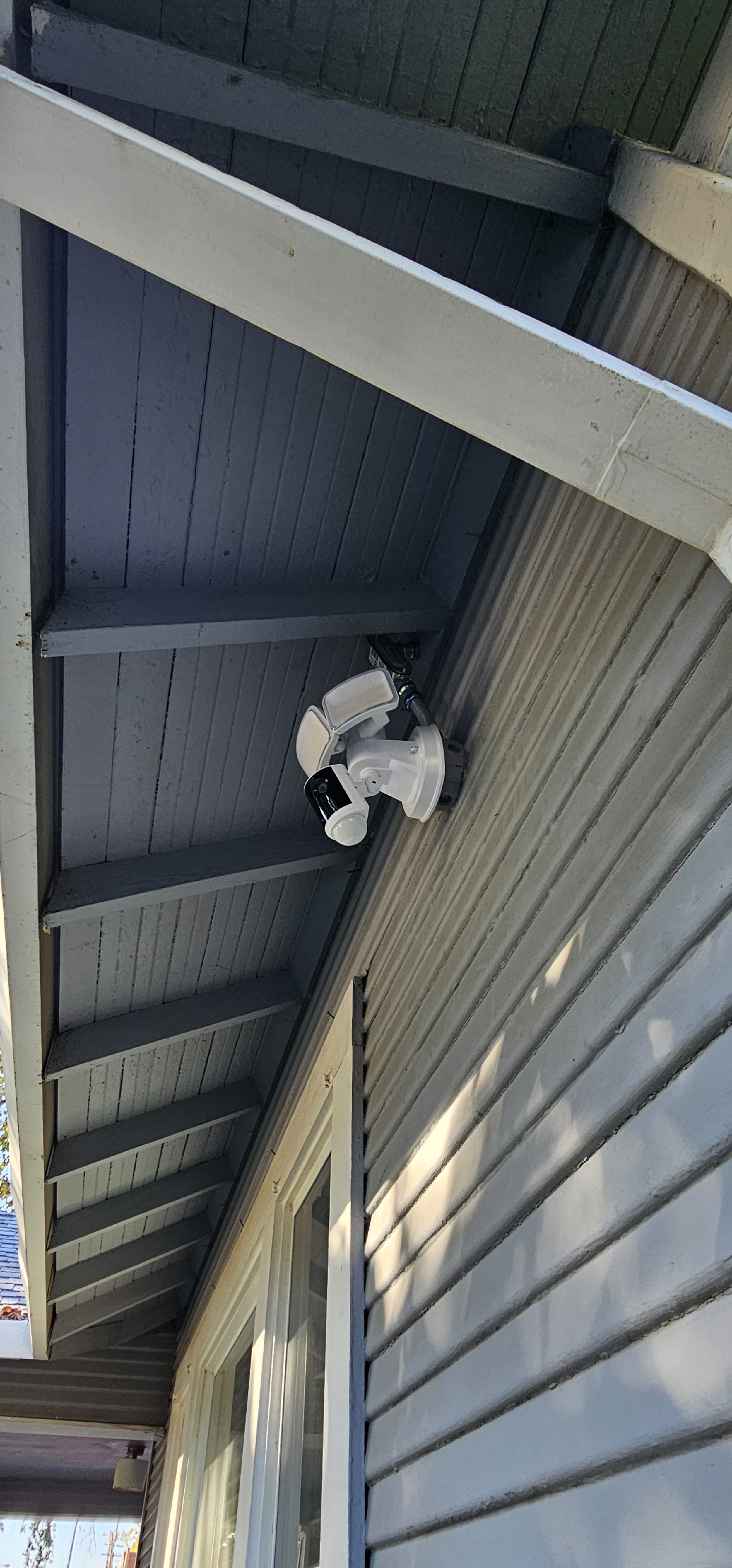 View of the underside of a house's roof, showing gray-painted wooden beams and siding, with a white security camera mounted under the eaves.