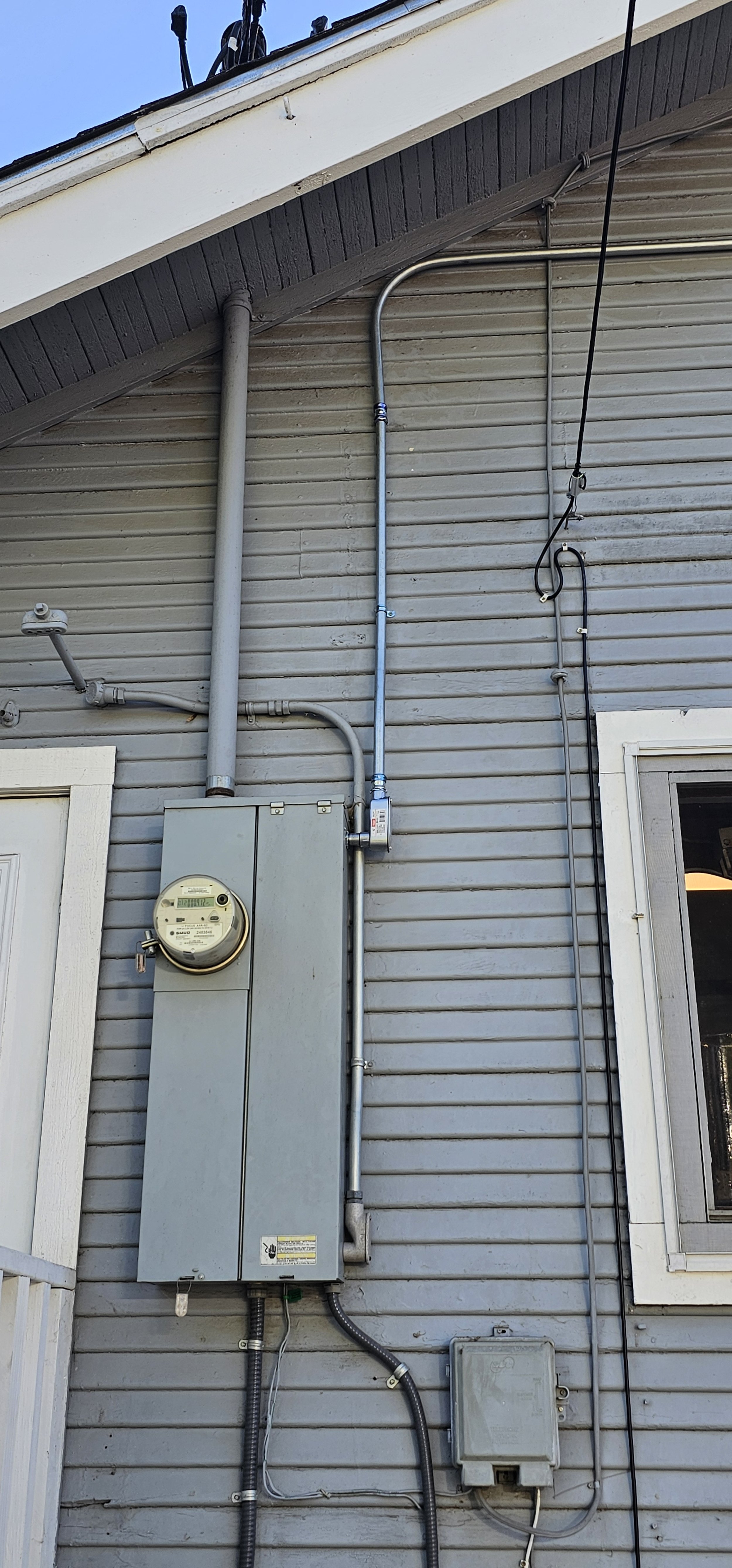 Close-up of house exterior showing electrical meter, electrical panel, conduit pipes, and electrical wiring on a gray wall.