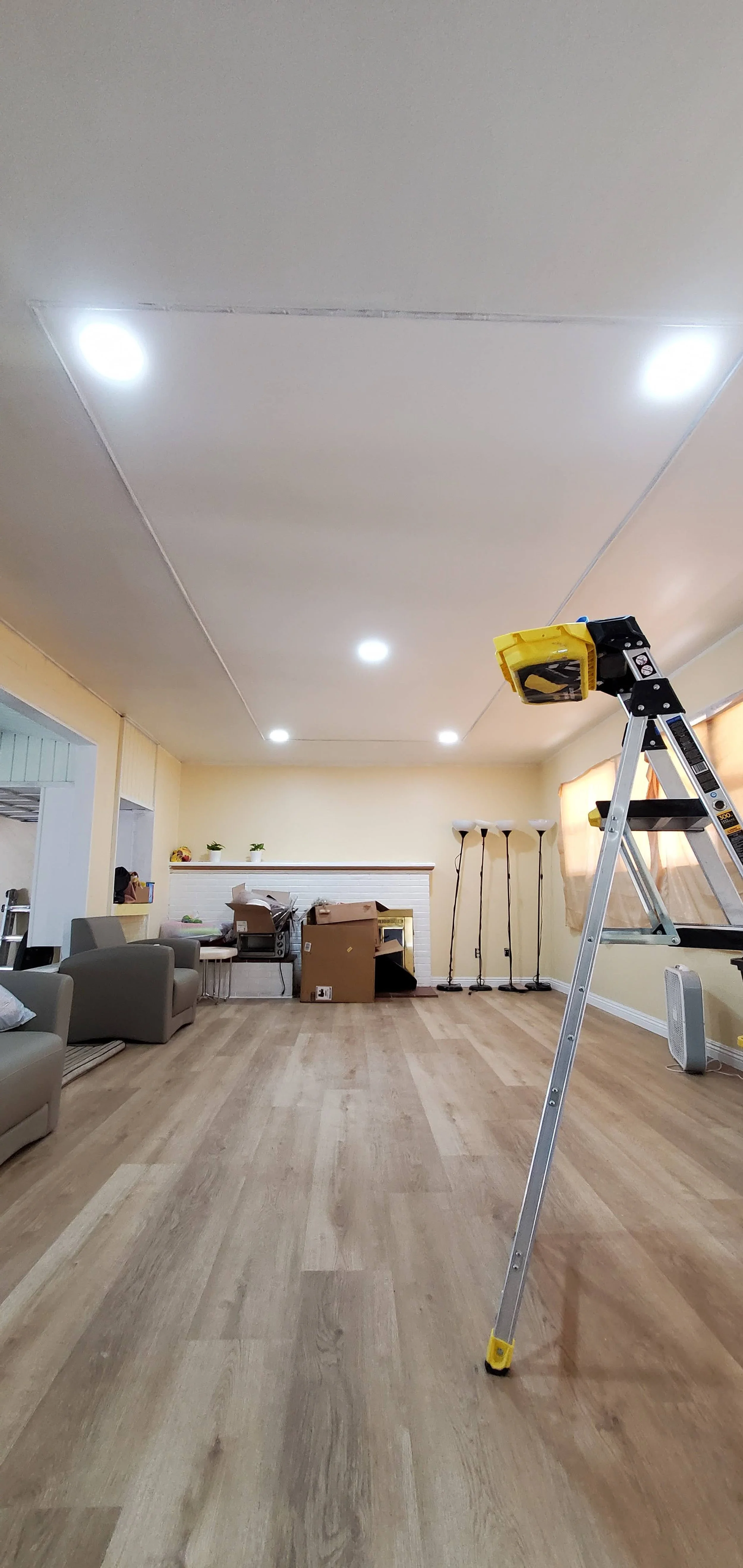 Living room with hardwood floors, a long white fireplace mantle, and a ladder with a yellow tool bag on top. There are chairs, boxes, and floor lamps. The ceiling has recessed lights.