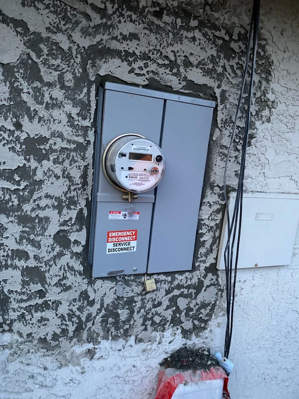 Electric meter box attached to a textured exterior wall with a red and white emergency disconnect sign, a padlock, and electrical cables running vertically, with debris and a white box on the ground.