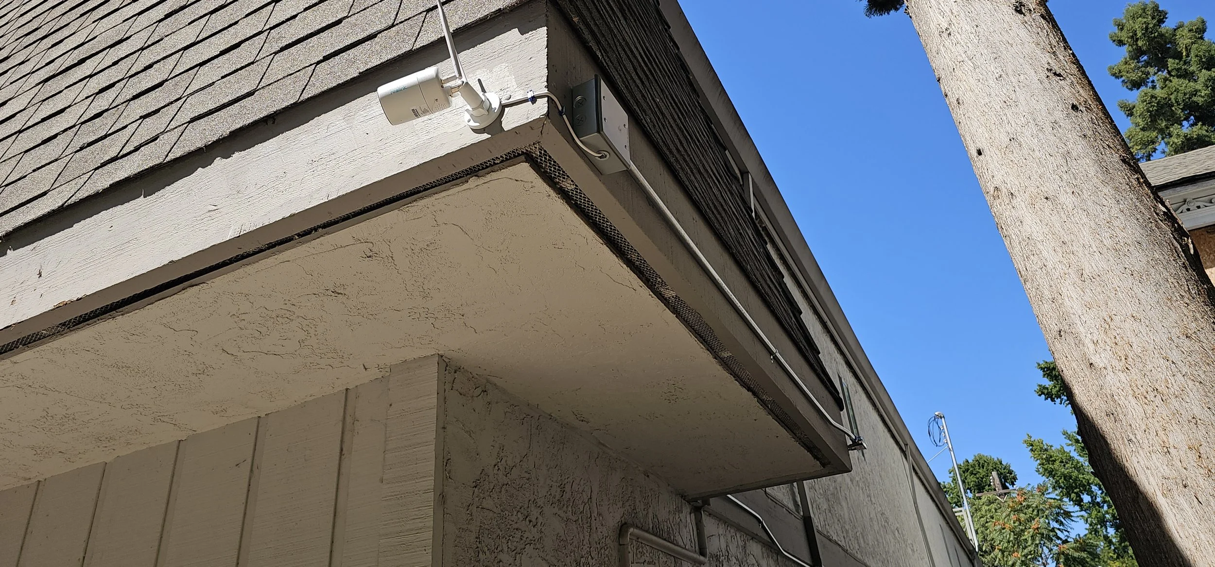 Close-up of the corner of a house's overhang showing a security camera, electrical box, cable conduit, and a tree trunk nearby, with a clear blue sky in the background.