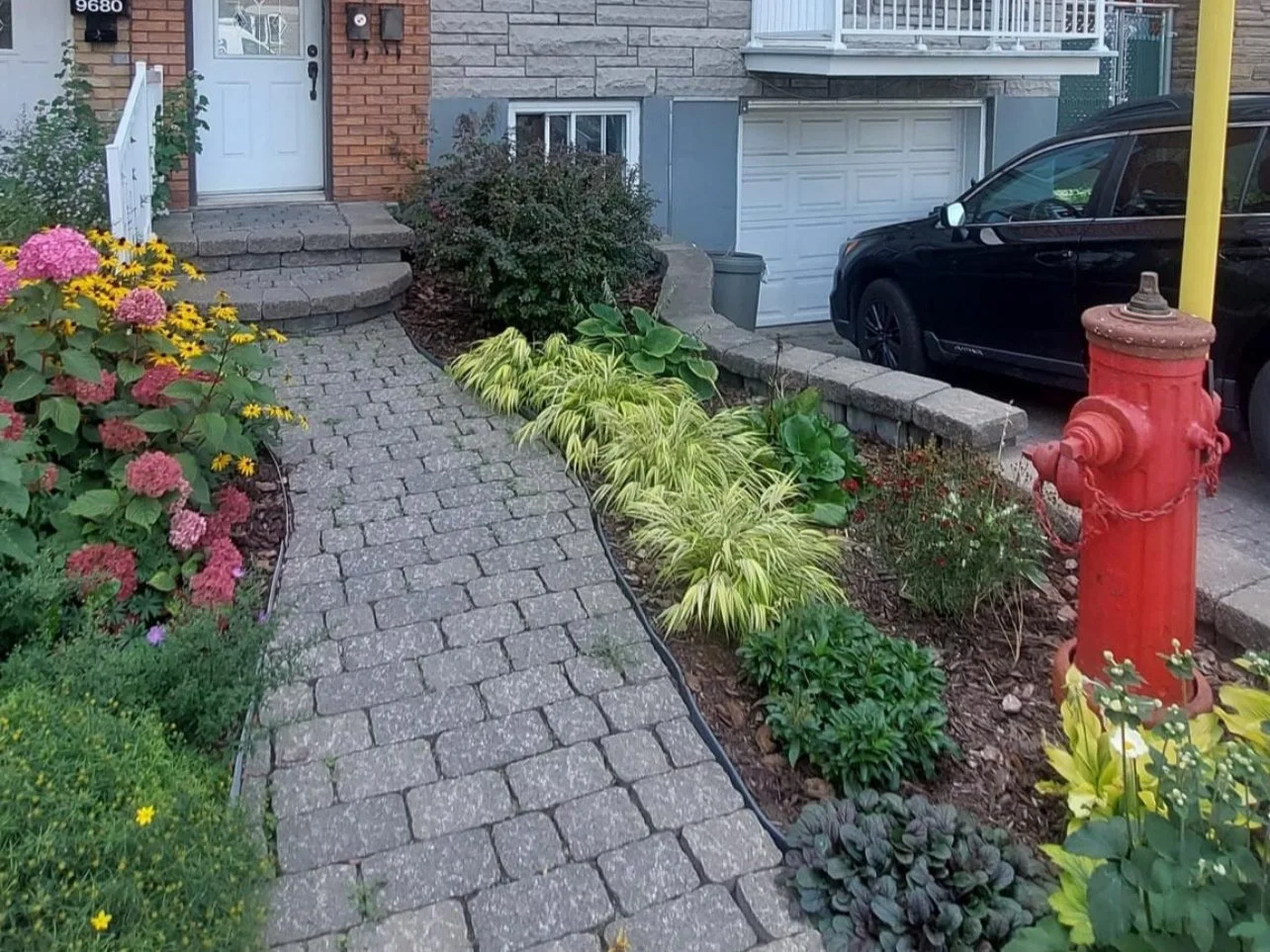 Entrée de maison avec jardin fleuri, chemin en pavés, bouche d'incendie rouge et voiture noire garée.