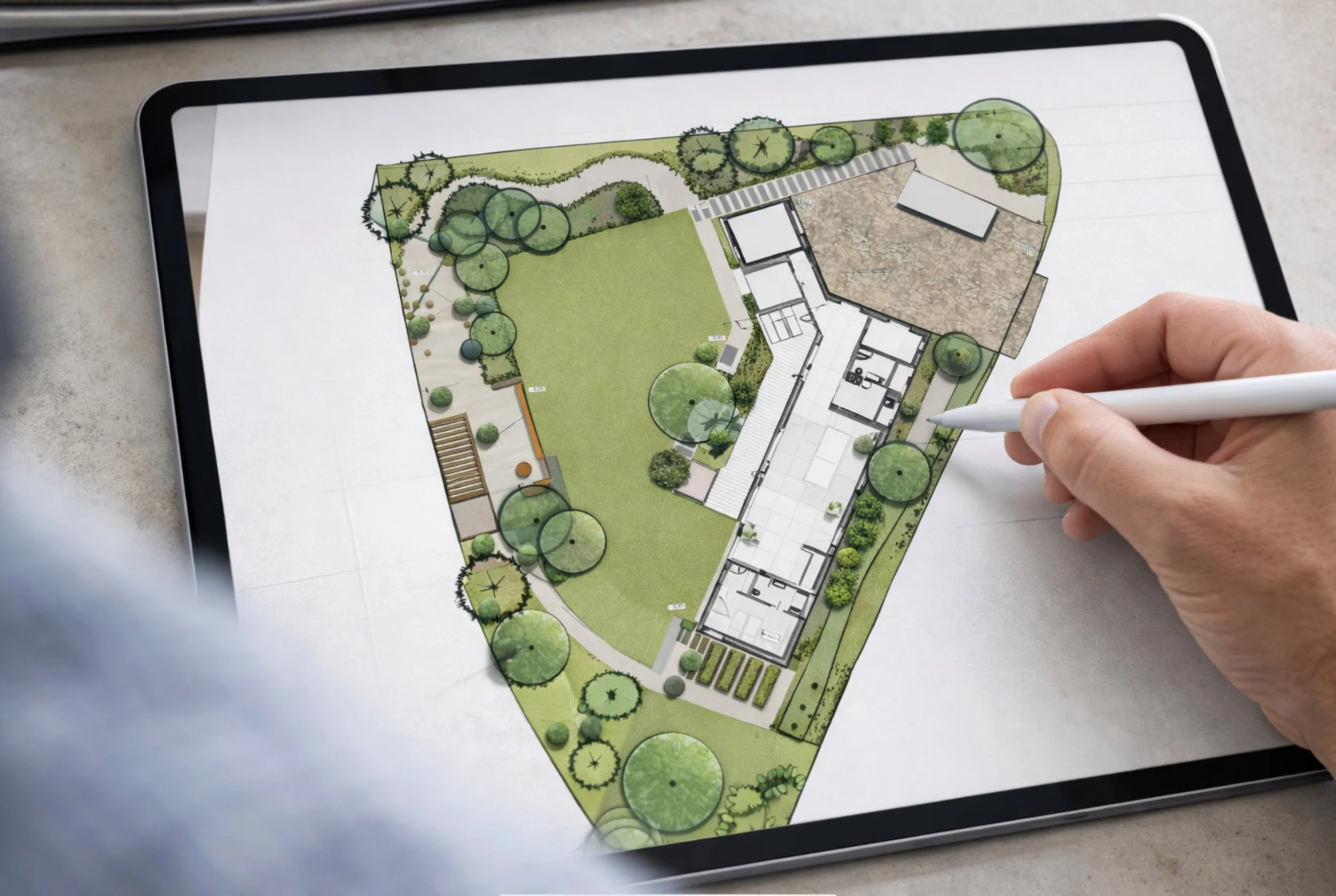 Plan d'aménagement paysager affiché sur une tablette, avec un dessin de jardin comprenant des arbres, une pelouse, un patio et des zones de plantation.