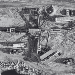 Black and white aerial photo of a mine site with heavy machinery, cranes, and vehicles working on the earth.
