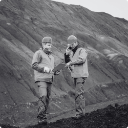 Two mine site workers in hish visibility PPE and hard hats stand outdoors on a mine site. They are looking at a document together.