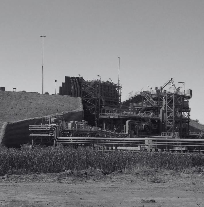 A large industrial plant with multiple structures and pipes, situated on a hillside with a grassy foreground.