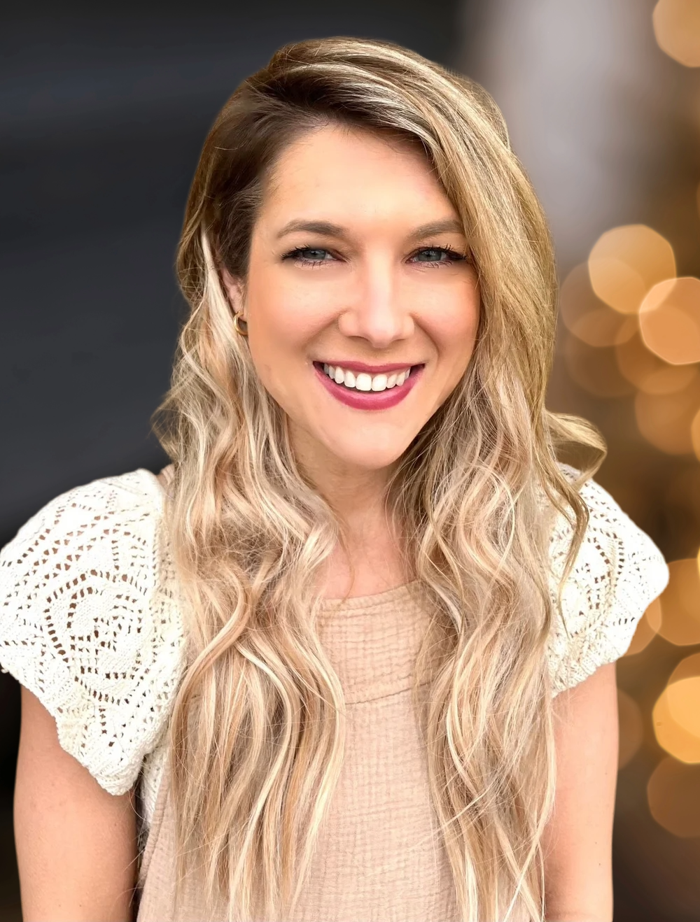 A smiling woman with long, wavy blonde hair, wearing makeup and a light-colored top.