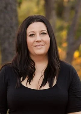 A woman with long wavy brown hair and blue eyes sitting cross-legged on the floor in front of a beige sofa, wearing a white t-shirt and blue jeans, smiling at the camera in a modern room with dark green walls and a large green potted plant to her right.