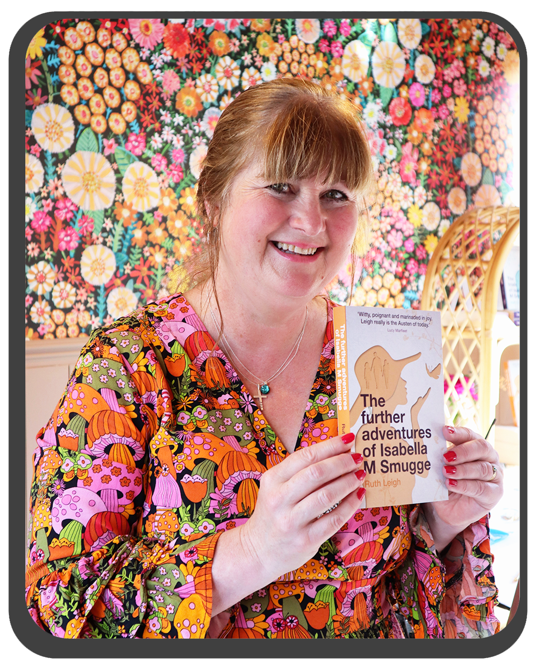 Author Ruth Leigh, wearing a colourful floral-patterned blouse and a cross necklace, holding a book titled 'The Further Adventures of Isabella M. Smugge' by Ruth Leigh, standing in front of a vibrant, floral tapestry background.
