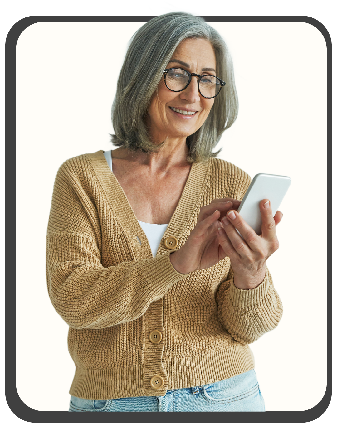 An older woman with gray hair and glasses, smiling and looking at her smartphone, wearing a tan cardigan and jeans.