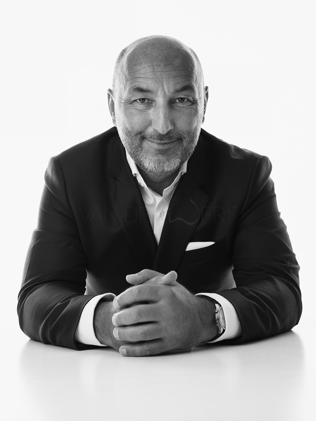 Schwarz-weiß Porträt eines lachenden, älteren, bärtigen Mannes in formellem Anzug mit weißen Hemd, sitzt mit verschränkten Händen auf einem Tisch.