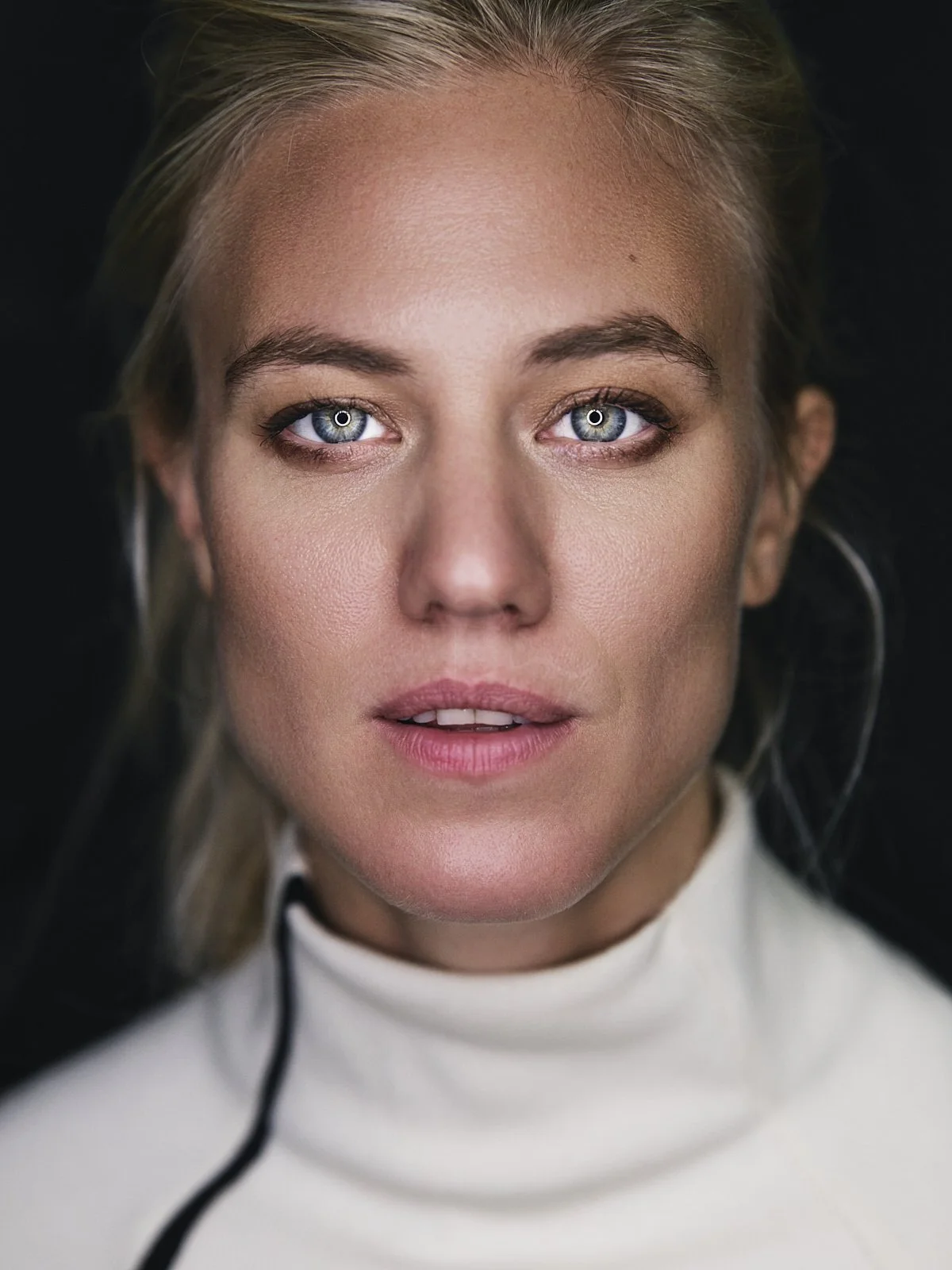 Nahaufnahme eines Mädchens mit blauen Augen, hellem Haar und einem weißen Sportoberteil auf schwarzem Hintergrund.