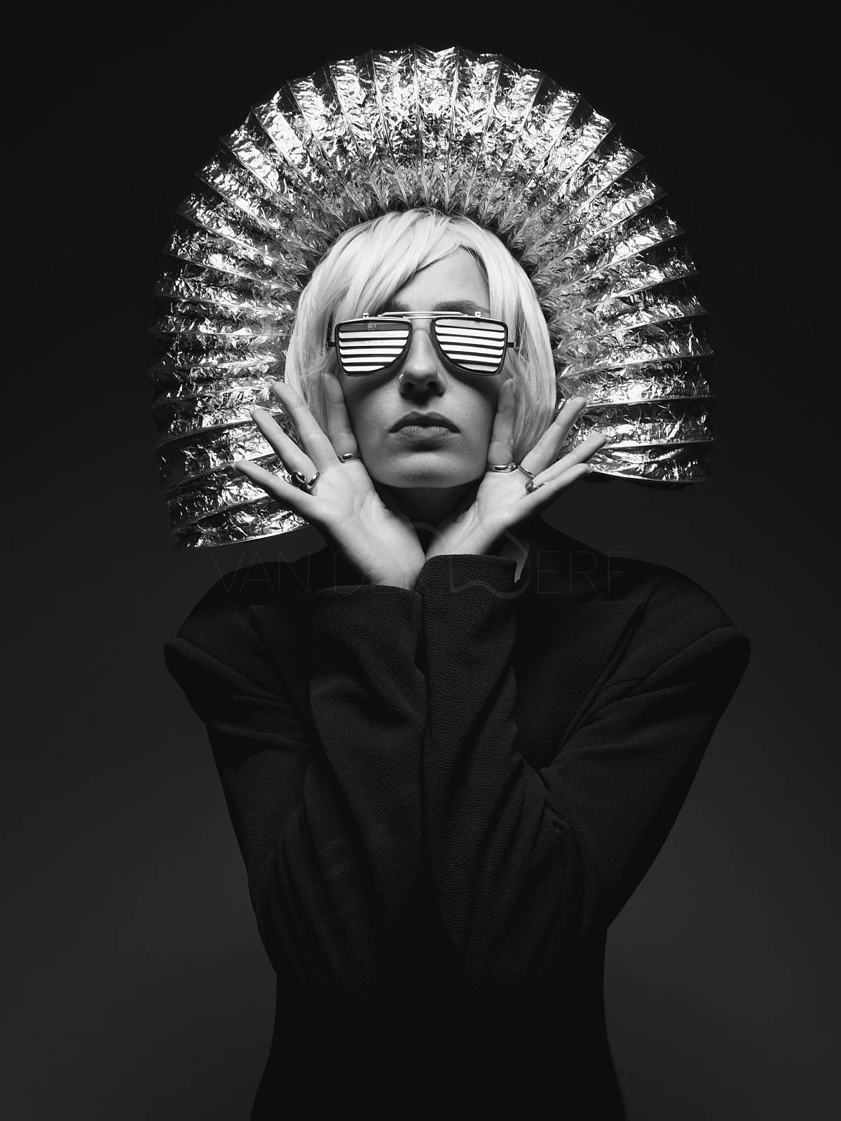 Schwarzweißer Porträt einer Frau mit futuristischem Design. Sie trägt große, reflektierende Sonnenbrillen mit horizontalen Linien, eine auffällige silberne Kopfbedeckung, schwarze Kleidung und mehrere Ringe. Die Pose ist stilistisch ausgeprägt, um ei