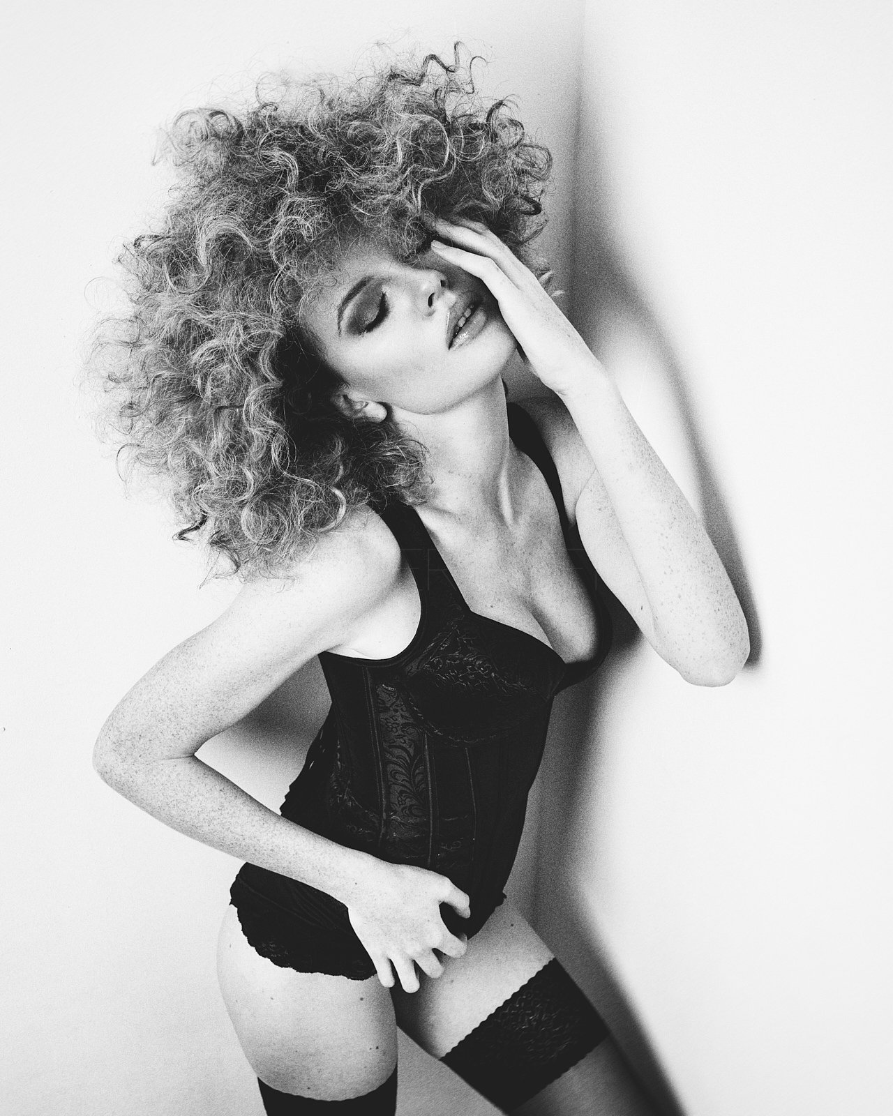 Schwarz-weiß-Fotografie einer Frau mit lockigem Haar, die an der Wand lehnt und die Hand an das Gesicht legt, trägt schwarzen BH, Shorts und Strümpfe.