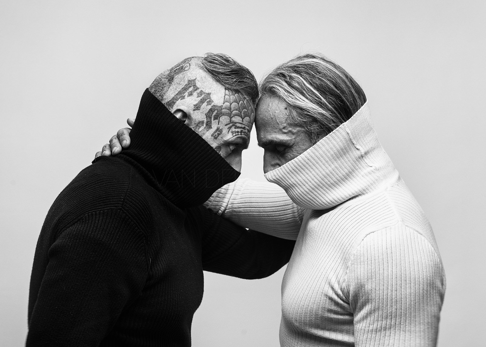 Zwei Männer stehen sich mit niedergeschlagenen Stirn gegenüber, tragen hochgekrempelte Pullover und haben ihre Arme umeinander gelegt, in einer tiefen, emotionalen Geste.