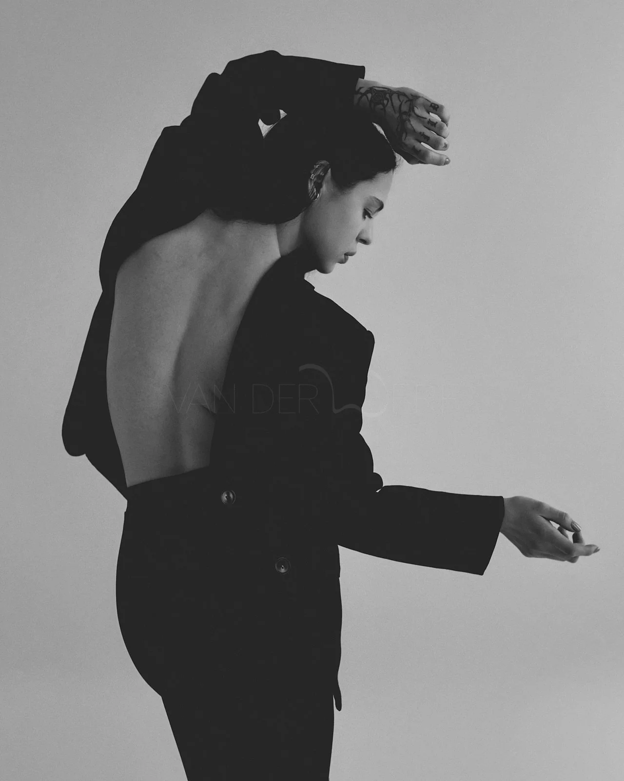 Schwarz-weiße Fotografie einer Frau mit offenem Rücken, die ihre Arme über dem Kopf hält, mit einem Tattoo auf ihrer Hand. Sie trägt einen schwarzen Anzug, lässt ihre Haare offen und schaut nach unten.