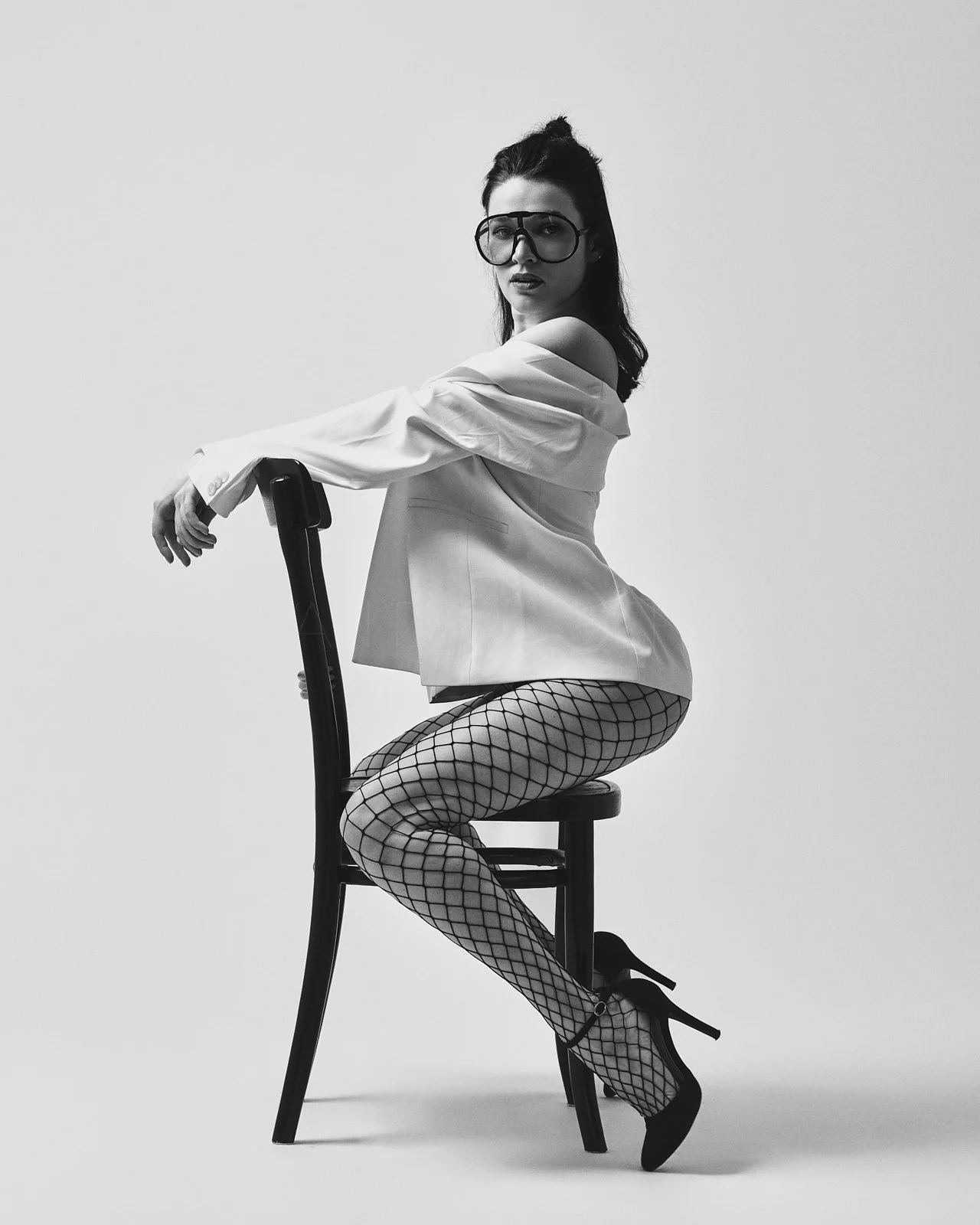 Schwarz-weiß-Fotografie einer Frau auf einem Stuhl sitzend, mit Netzstrümpfen, High Heels, off-Shoulder-Bluse und großen Brillen, frontal gesehen, mit ernstem Gesichtsausdruck.