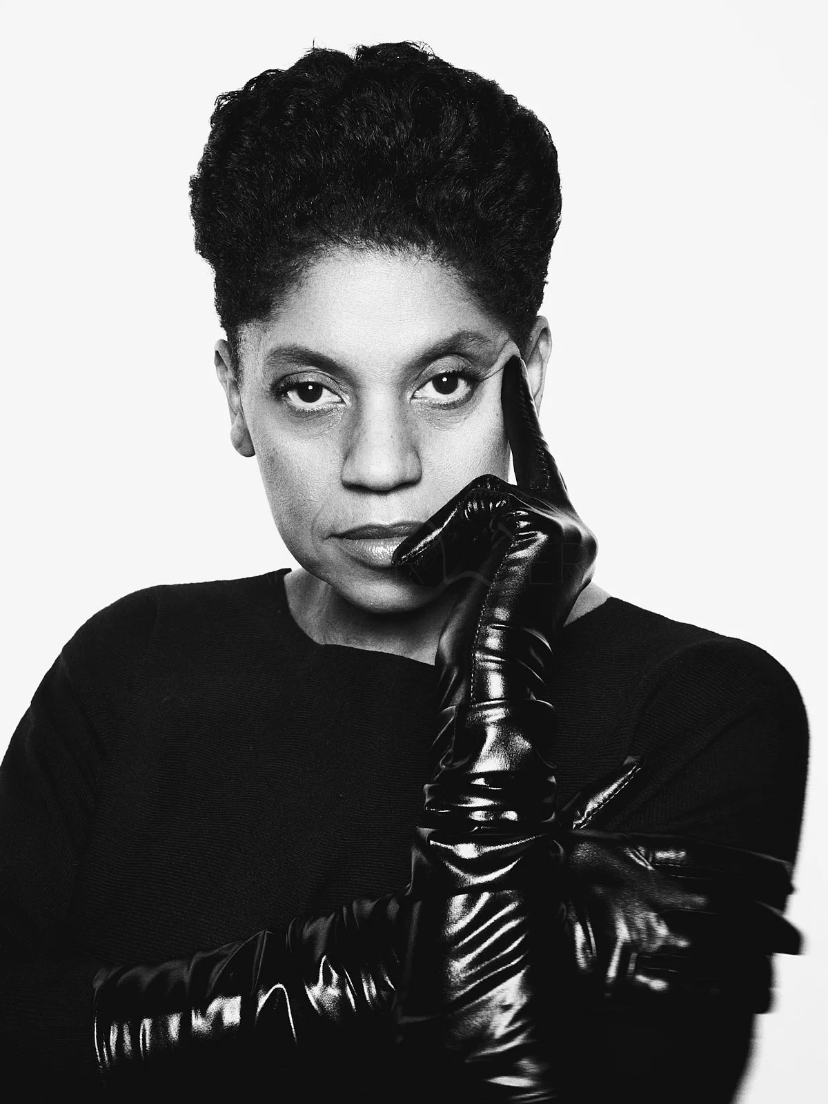 Schwarz-Weiß-Foto einer schwarzen Frau mit kurzes, volles Haar, die eine schwarze Lederhandschuh trägt und eine Hand an ihr Gesicht legt. Blick direkt in die Kamera.