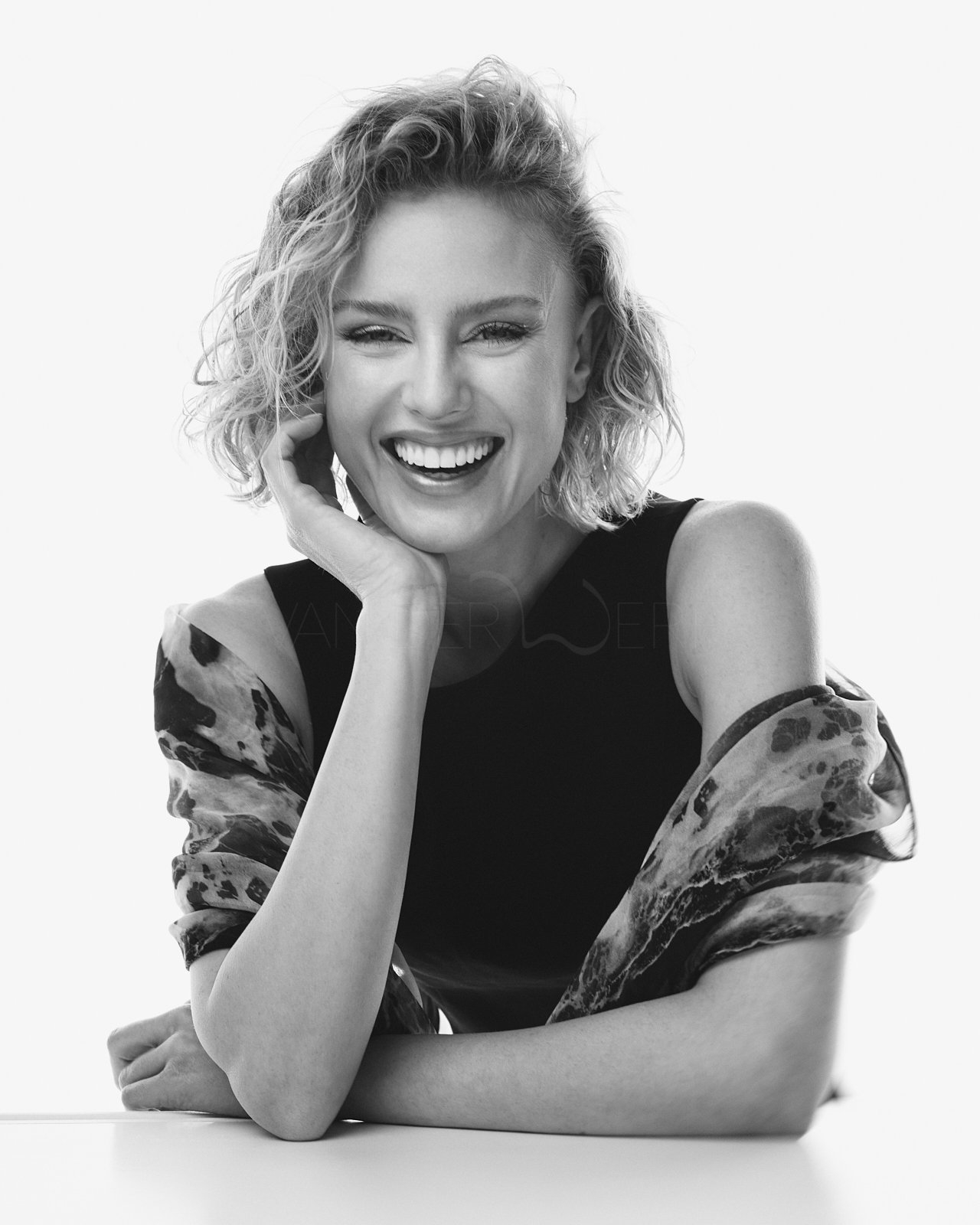 Ein lachendes, blondes, curly Haar Mädchen mit einem schwarzen Oberteil und einer durchsichtigen, gemusterten Jacke, die von der Schulter fallen gelassen ist, in Schwarz-Weiß-Fotografie.