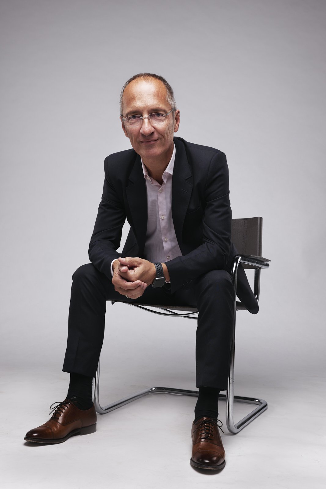 Ein Mann in Anzug sitzt auf einem schwarzen Stuhl vor hellgrauem Hintergrund, schaut zur Kamera und lächelt leicht.