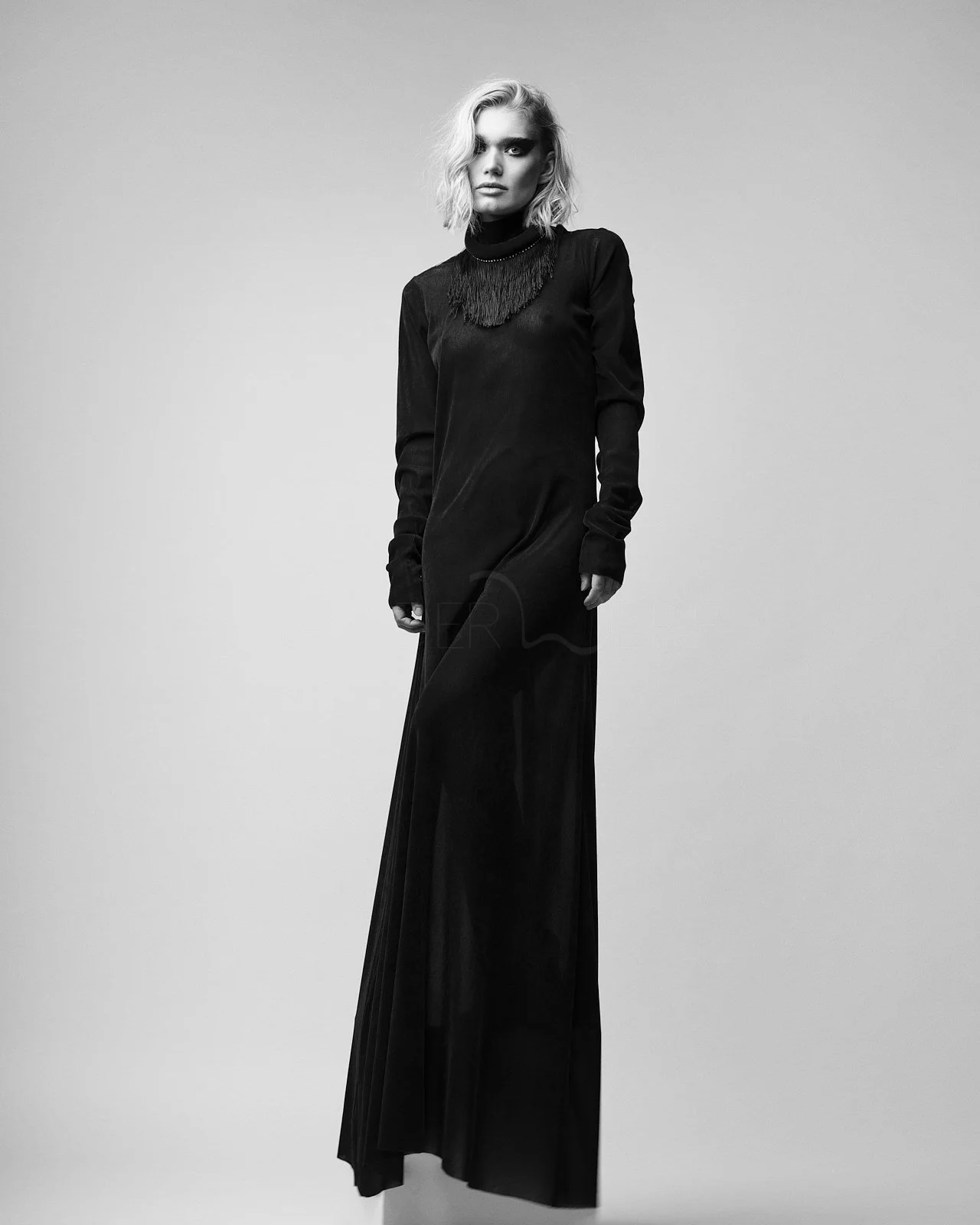 Schwarz-weiß-Foto einer Frau in einem langen schwarzen Kleid mit Fransen-Halskette, vor hellem Hintergrund.