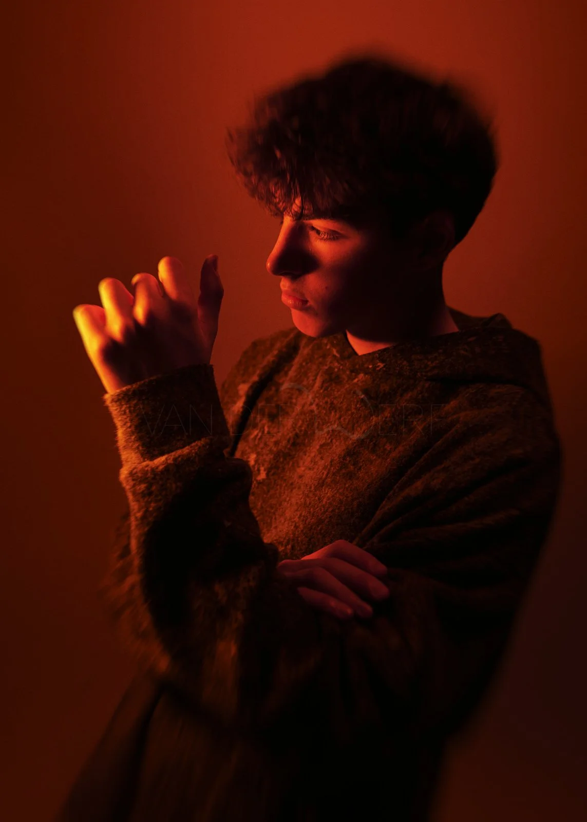 Junger Mann mit dunklem Haar und geschlossenen Augen, der in einem warmen, rot-orangen Licht angeleuchtet wird, trägt eine braune, weiche Jacke.